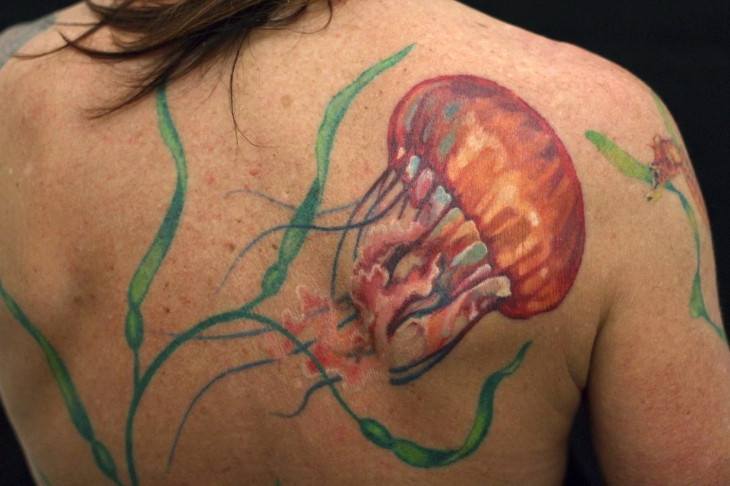 A realistic color jellyfish tattoo on the upper back features translucent pink and orange hues with flowing green tendrils.