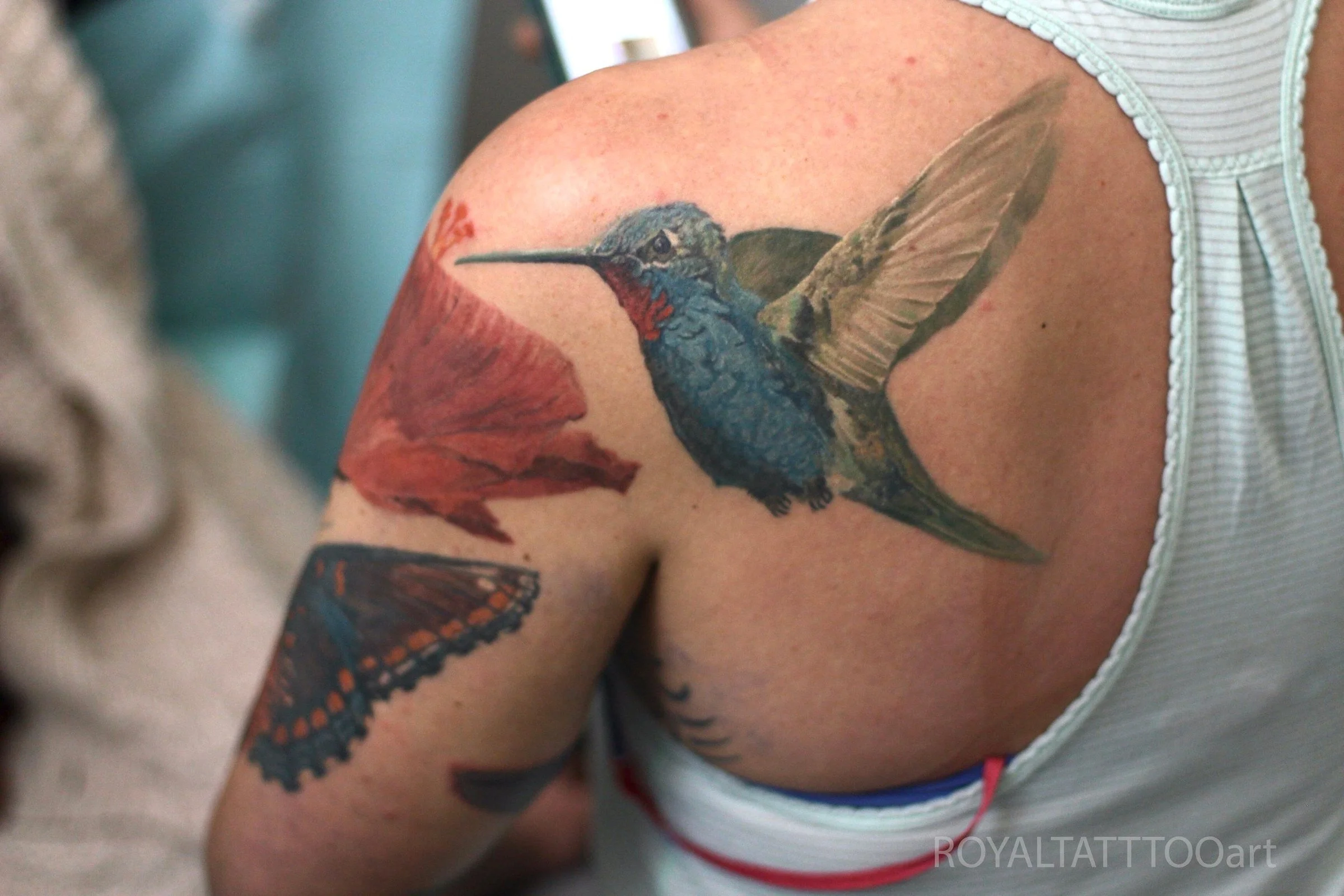 A color realism hummingbird tattoo on the shoulder blade shows the bird hovering near a flower, with soft shading and natural hues.