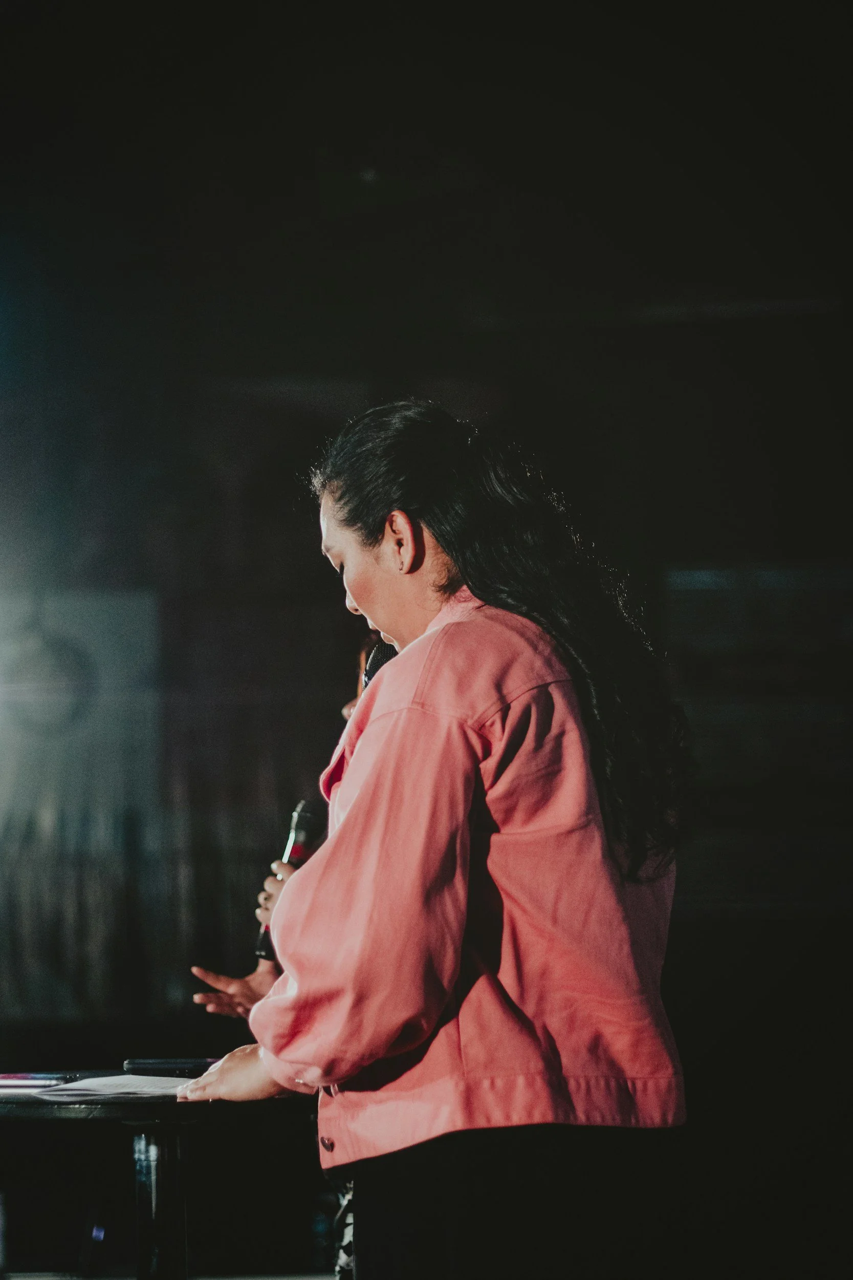 a person with long dark hair, wearing a coral pink jacket, holding a microphone, facing slightly away from the camera