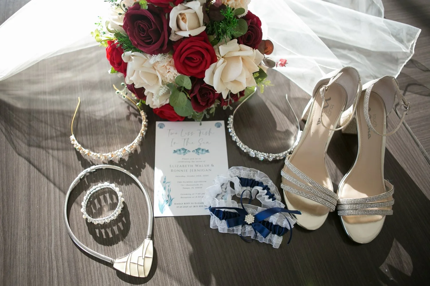 Bridal details with tiaras, necklace, garter, invitation, red and ivory rose bouquet and silver heels