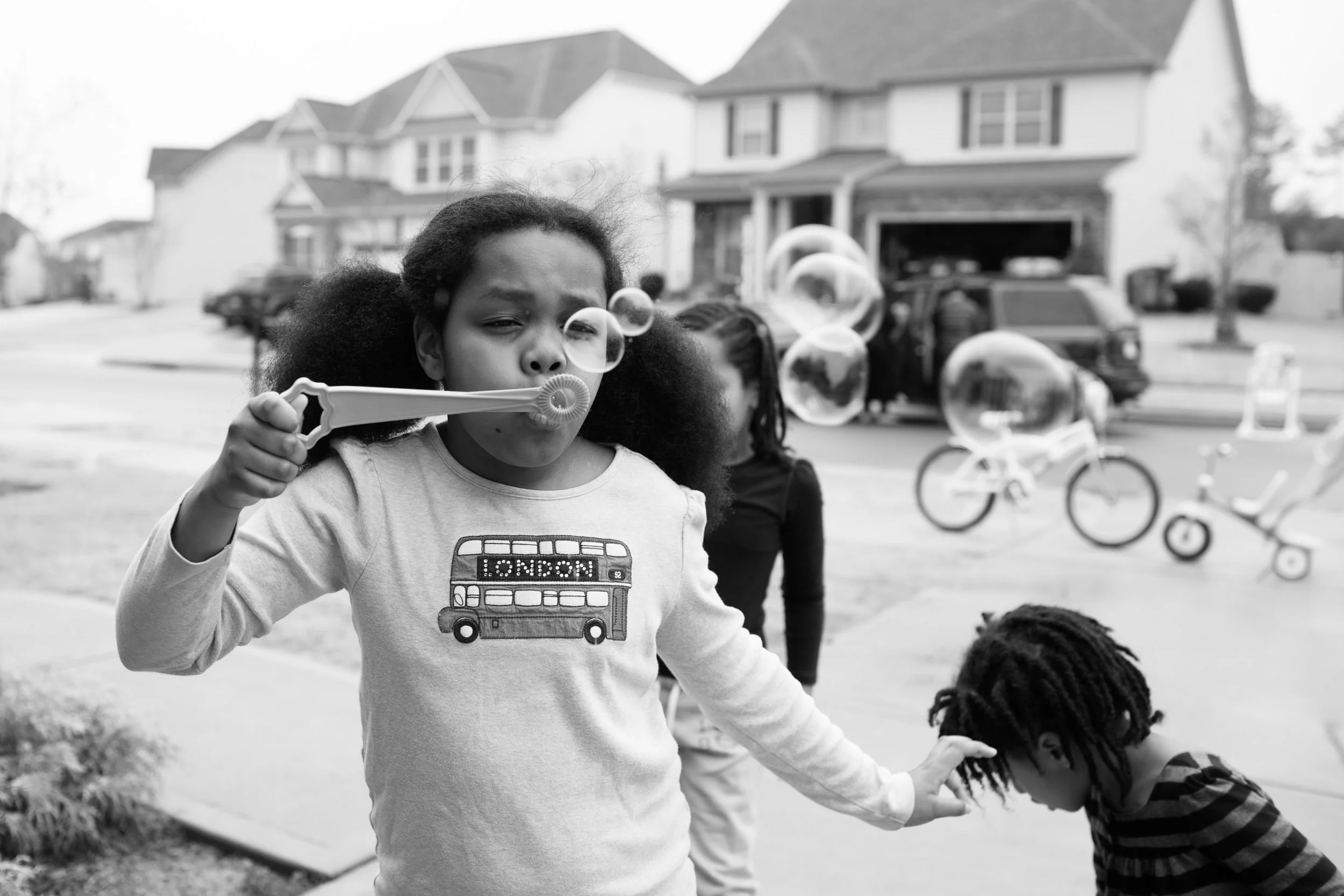 A young girl blowing bubbles outdoors in a residential neighborhood with children nearby, bicycles and houses in the background.