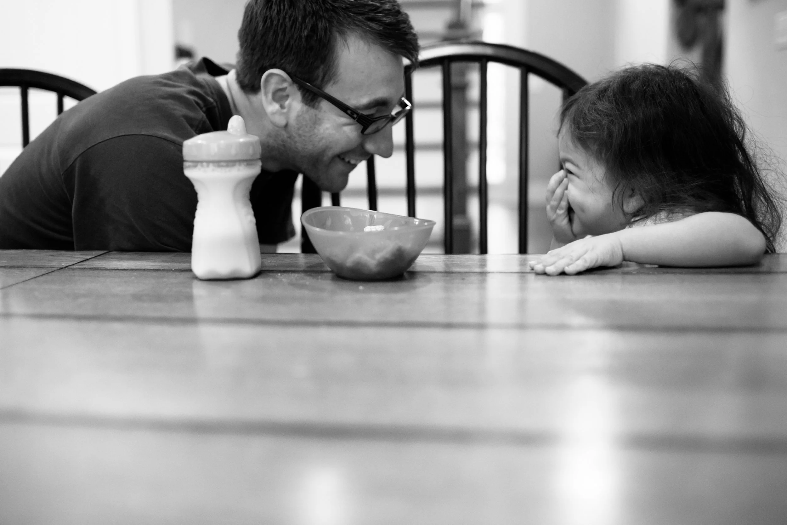 A man and a young girl sitting at a table, smiling and laughing at each other. The man is leaning forward with glasses on and the girl has her hand over her face, appearing playful. There is a bowl and a bottle on the table.