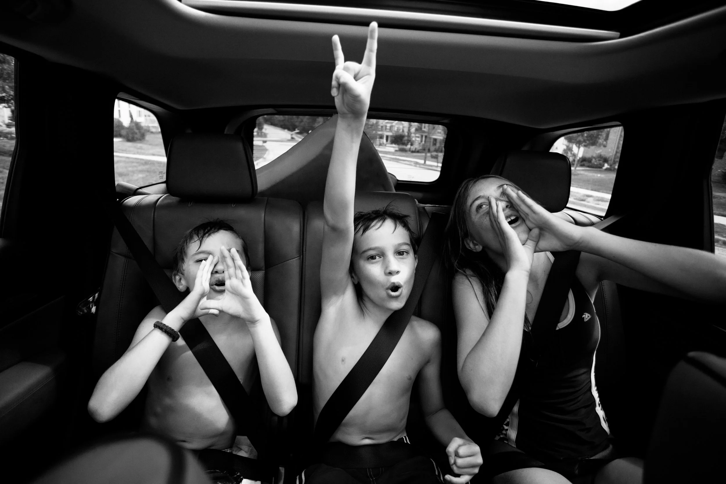 Three children in a car making playful gestures and faces, with one child raising an arm and another making a heart shape with his hands.