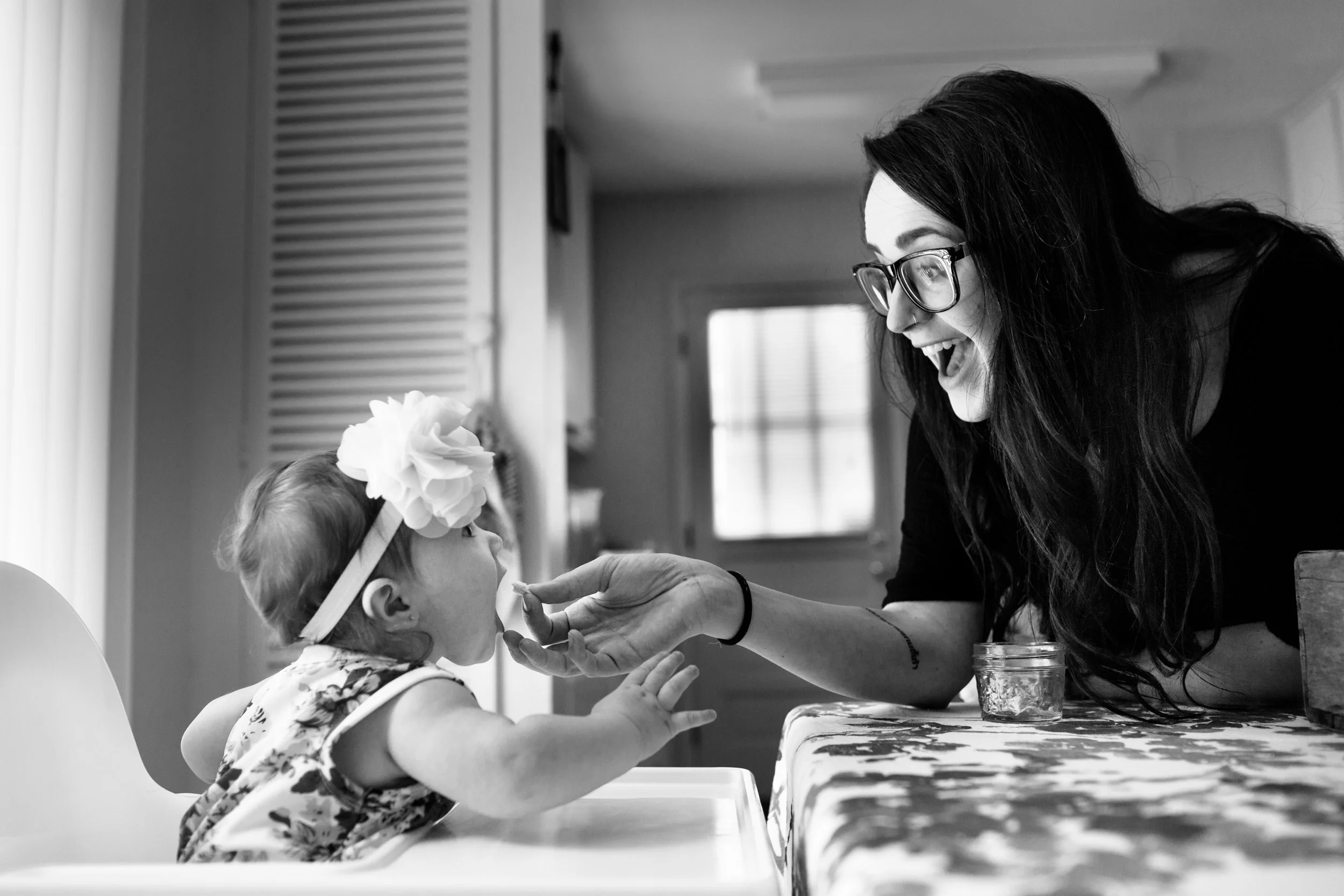 A woman with glasses and long dark hair is smiling and interacting with a young girl who is wearing a headband with a large flower. They are sitting at a table, engaged in a playful or nurturing activity in a home setting.