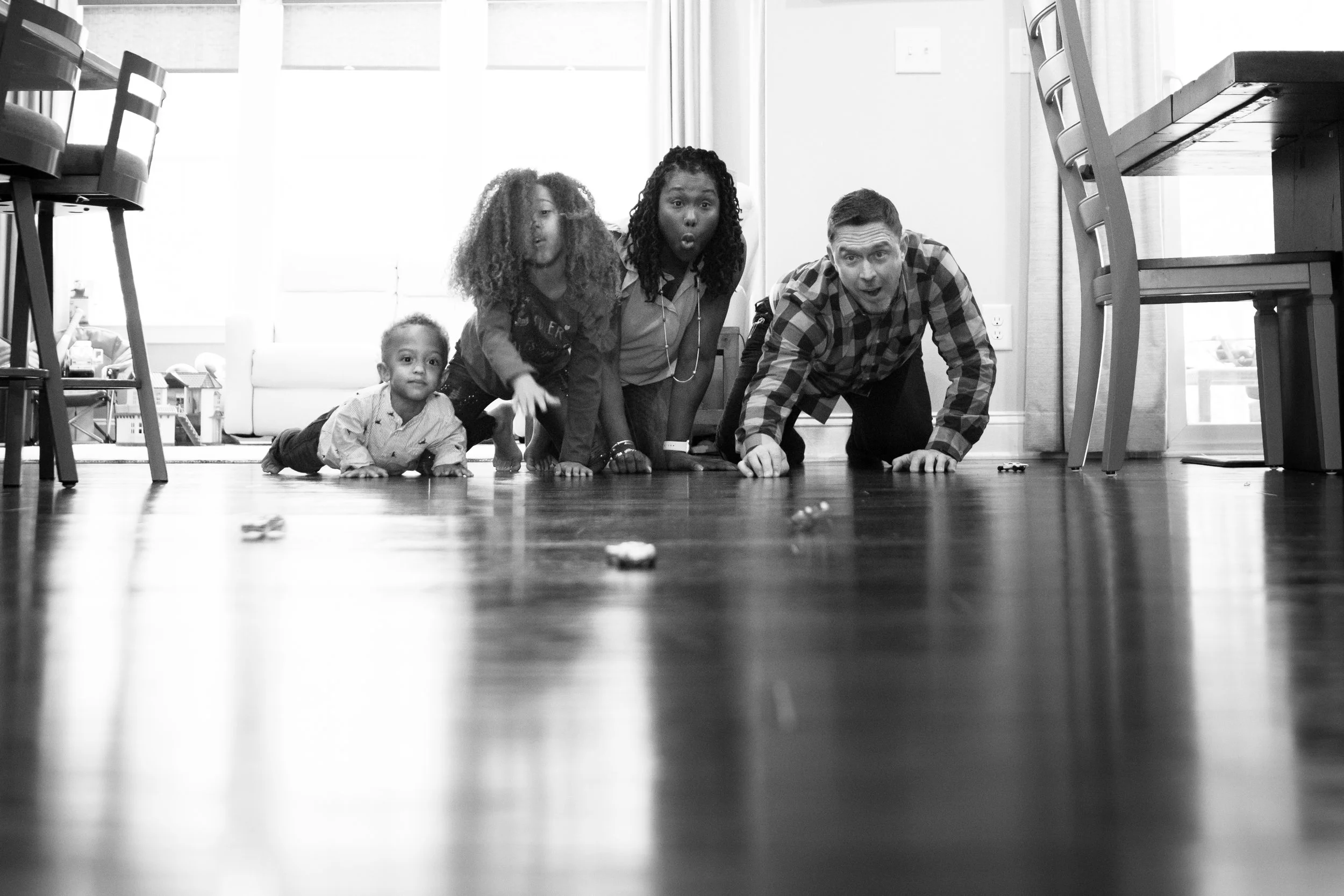 A man and three children crawling on the floor inside a house. The children are looking at the camera with surprised expressions, and the man is smiling. The floor is wooden, and there are chairs and a table in the background.