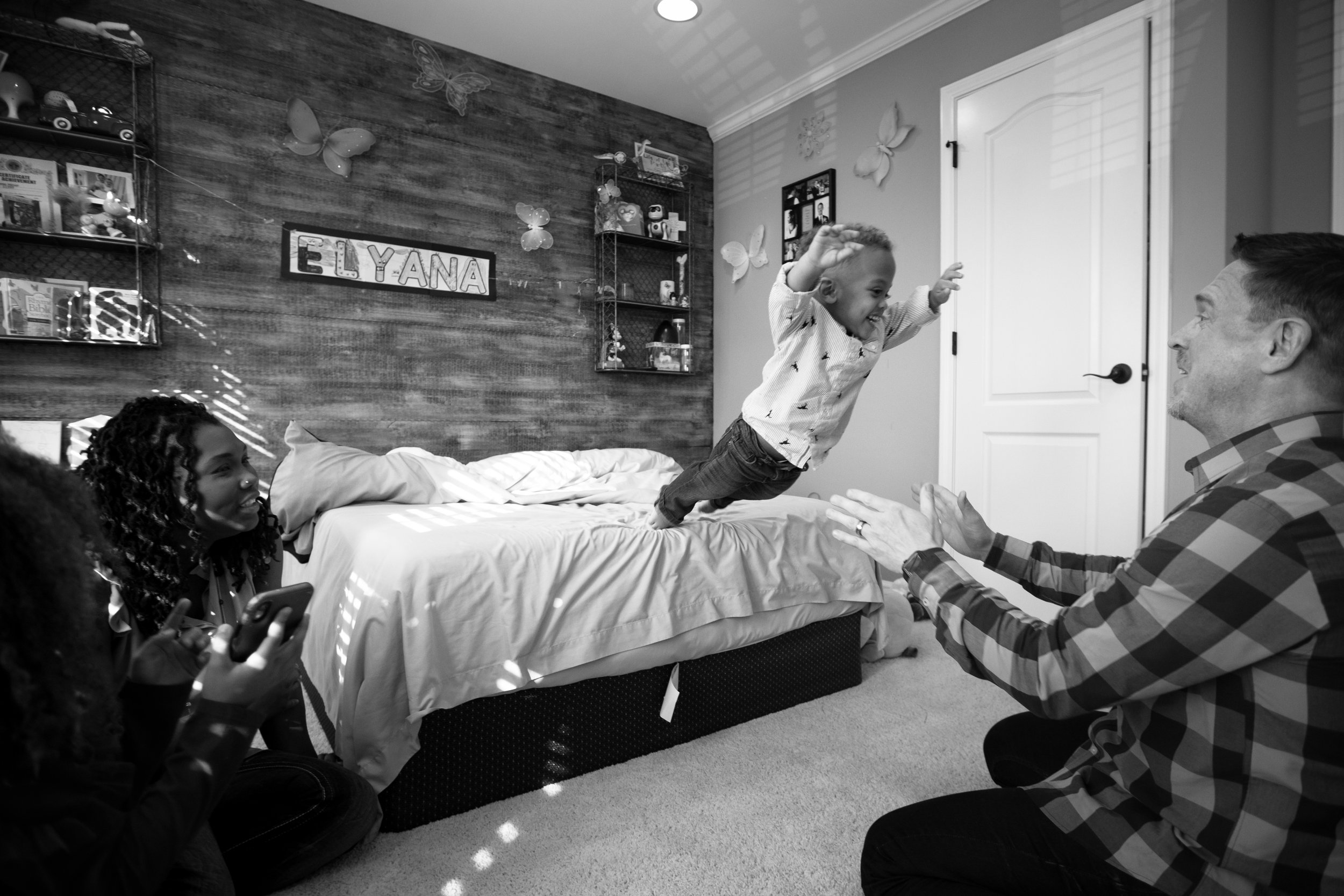 A young girl is jumping onto a bed, smiling with arms outstretched, while a man holds her hands. Another girl is sitting on the bed, watching. The room has butterfly decorations on the wall and a sign saying 'ELYANA'. The scene appears joyful and pla