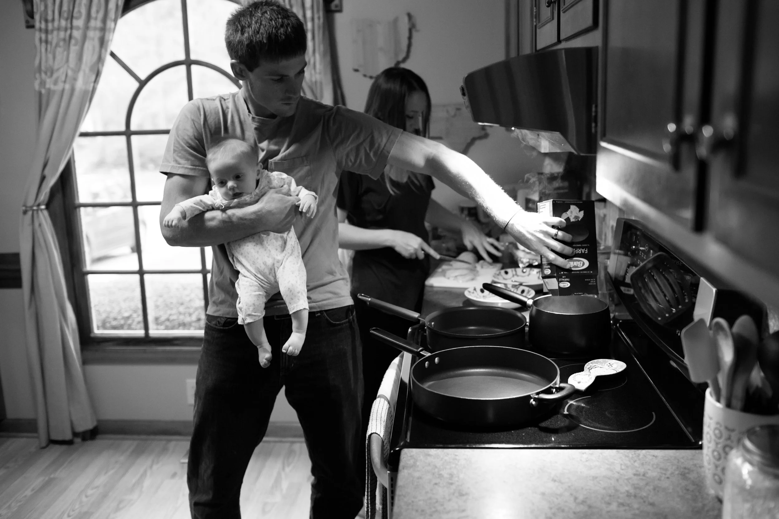 A man holding a baby while cooking in a kitchen. The man is reaching for a pot on the stove, and there is a woman in the background preparing food. The scene is in black and white.