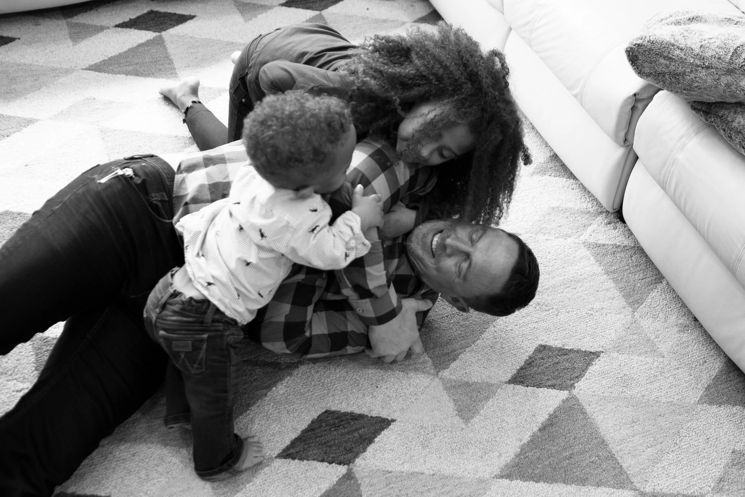 A woman with curly hair lying on a carpet with two children, playing together and smiling.