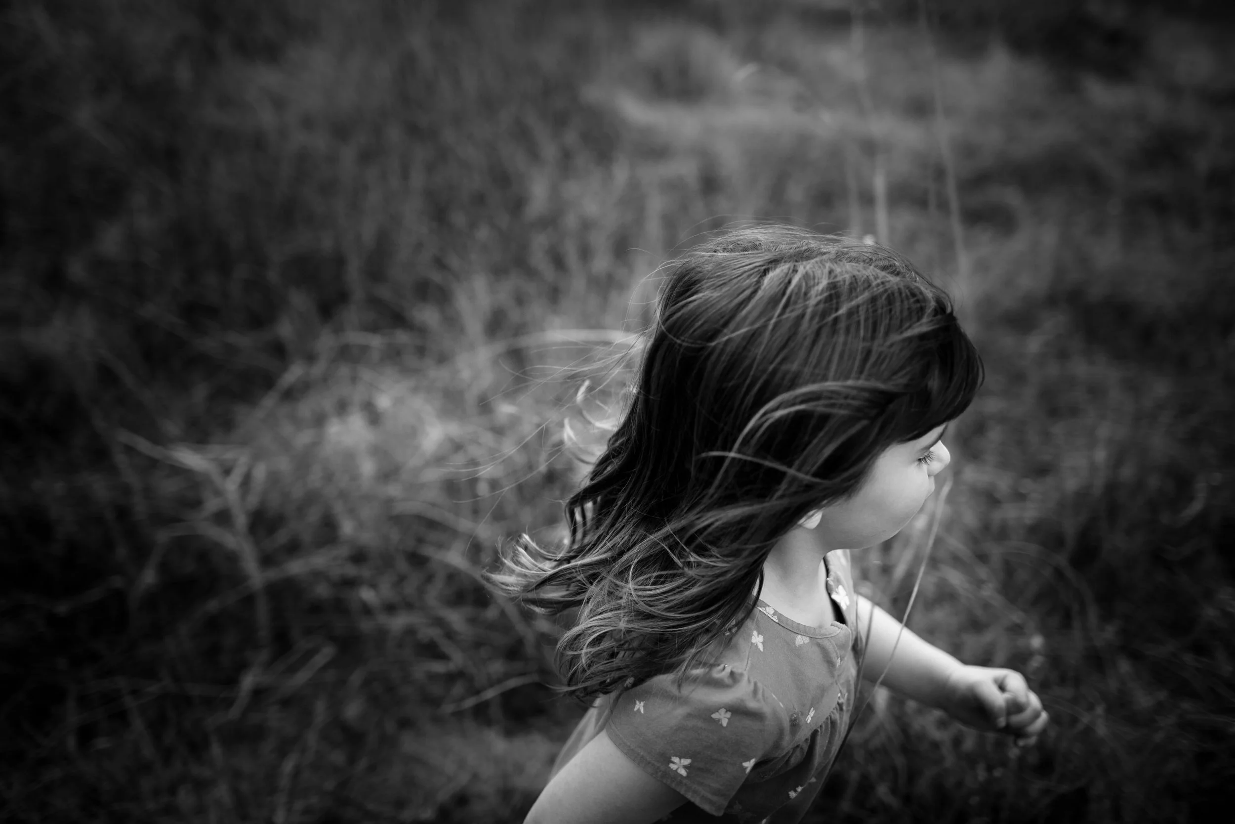 A black and white photo of a young girl with long hair, wearing a dress with a floral pattern, standing outdoors in a natural setting.