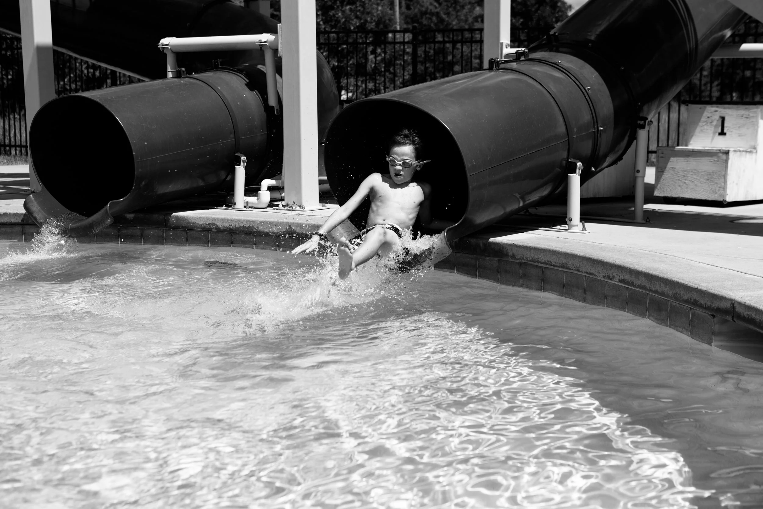 A young boy wearing goggles is emerging from a water slide at a swimming pool, with water splashing around him.