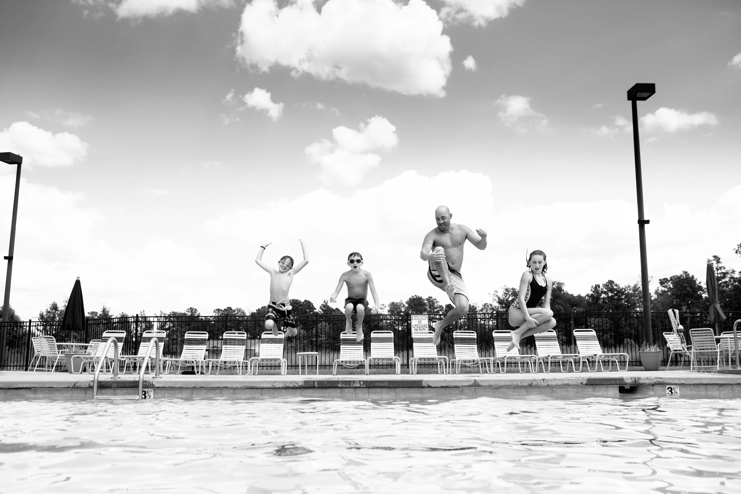 A black and white photo of four children and an adult jumping into a swimming pool from the pool's edge. The scene is outdoors with a cloudy sky and poolside chairs in the background.