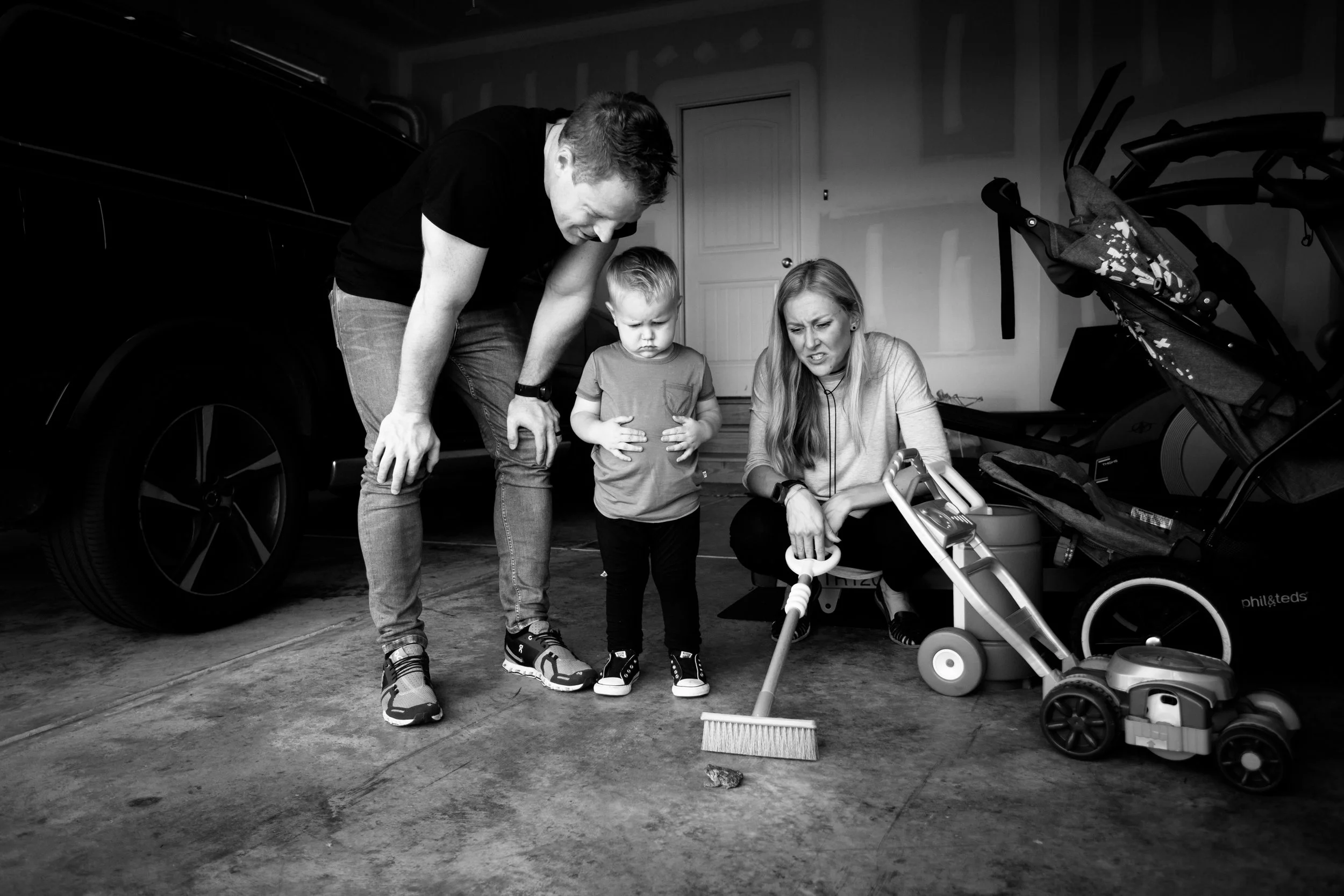 A family of three, consisting of a man, woman, and young boy, are kneeling on a garage floor, looking distressed at a small rock or object on the ground. The garage contains a bicycle, a stroller, and a car parked inside.