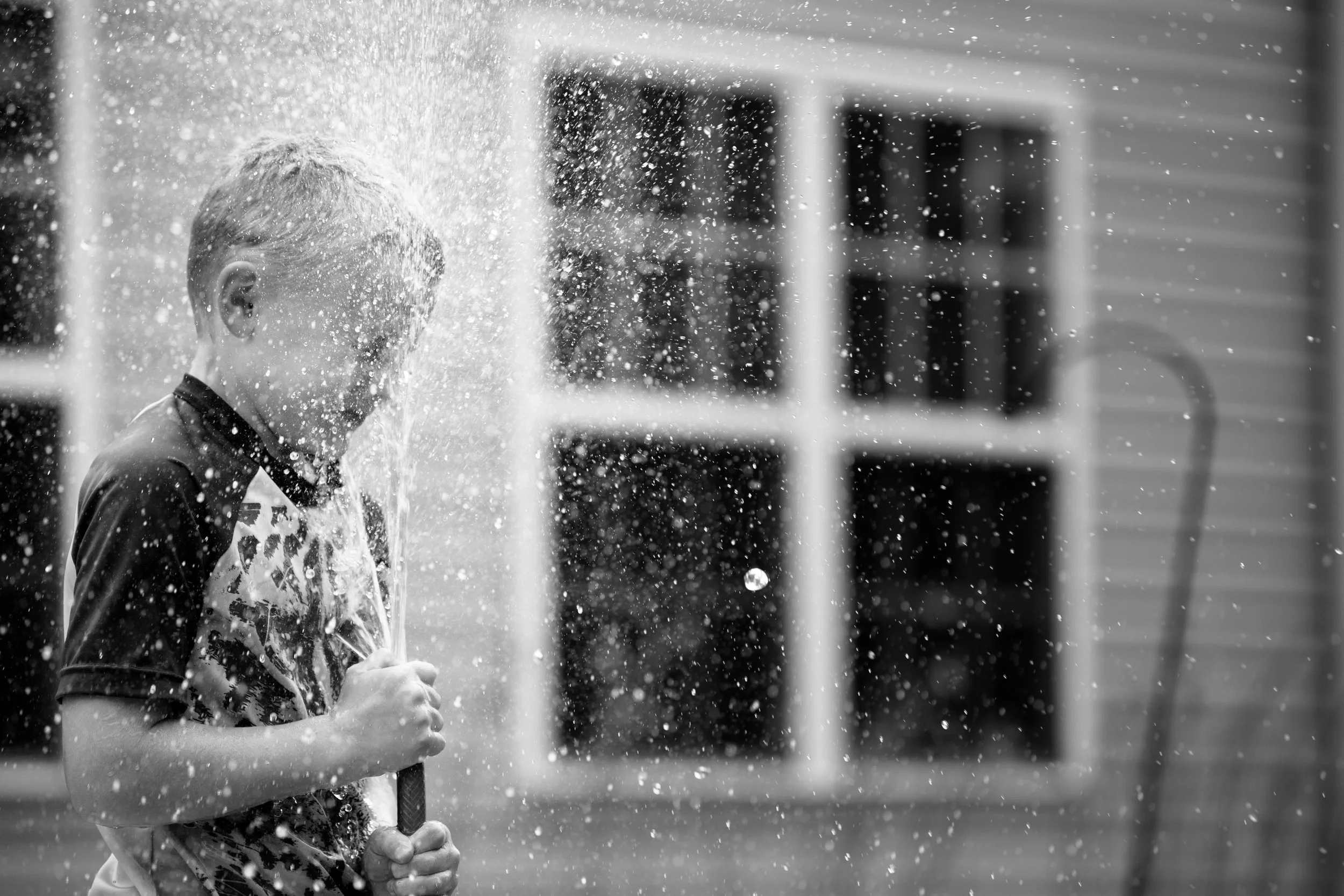 A young boy with wet hair and clothes is playing with a water spray, creating a splash and droplets in the air. The background features a house with a porch and a window.