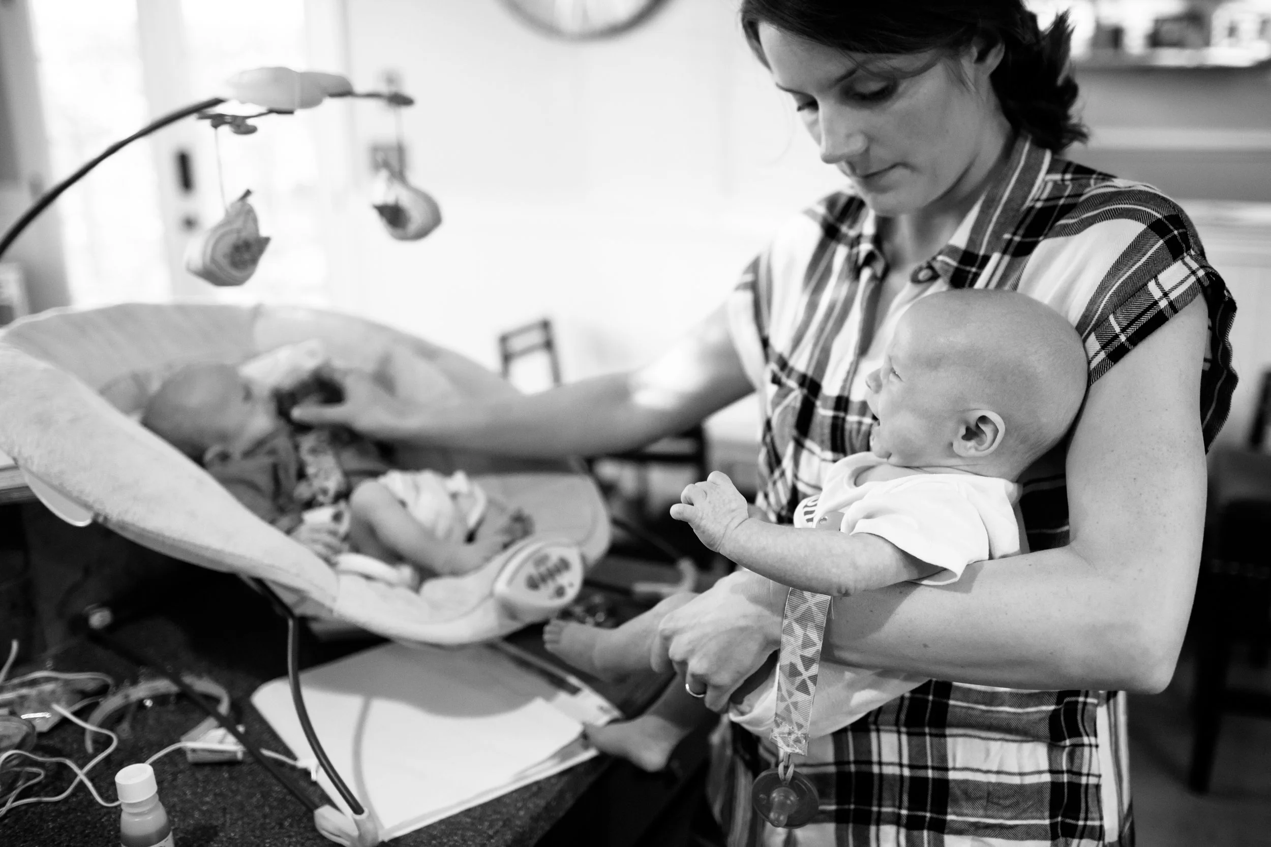 A woman in a plaid shirt holds a baby in a medical setting, with medical equipment and wires nearby, tending to the baby lying in a hospital bassinet.