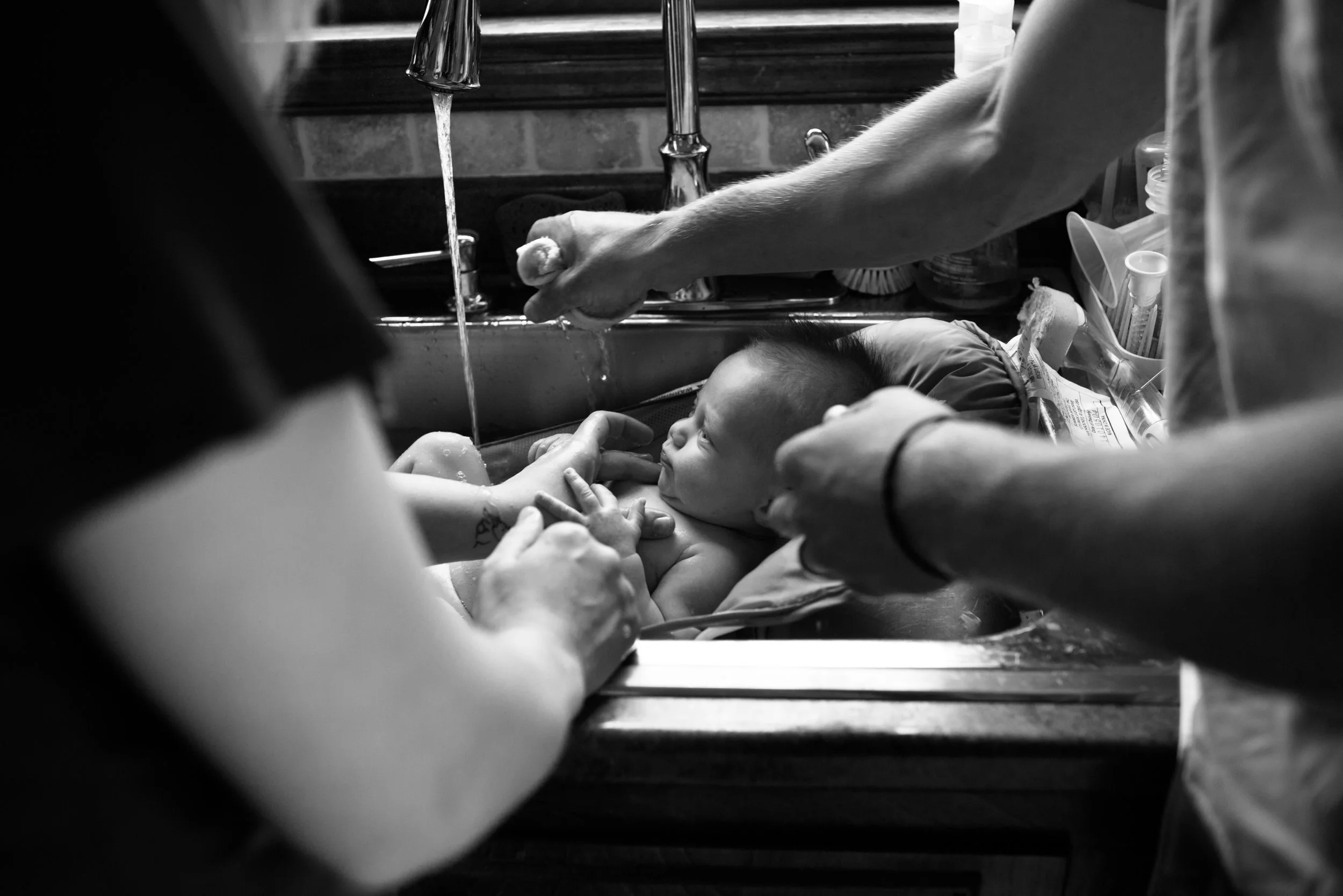 Black and white photo of a newborn baby being washed in a sink with the hands of two adults assisting, surrounded by various toiletries and supplies.