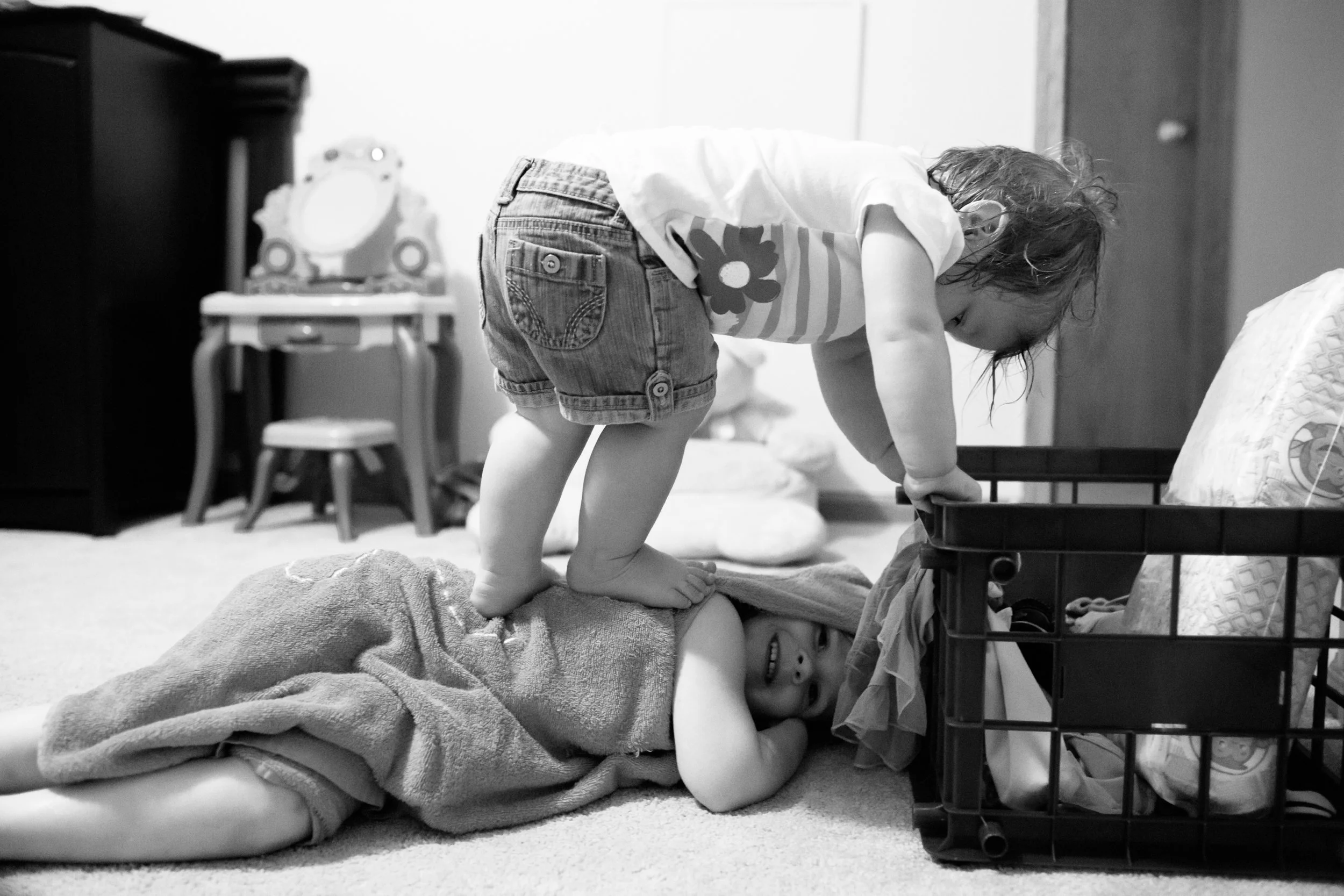 Two children playing, with one lying on a blanket on the floor and the other standing on them, inside a room. The standing child is leaning over and looking down at the child on the floor. The background includes a small table, a chair, and some toys