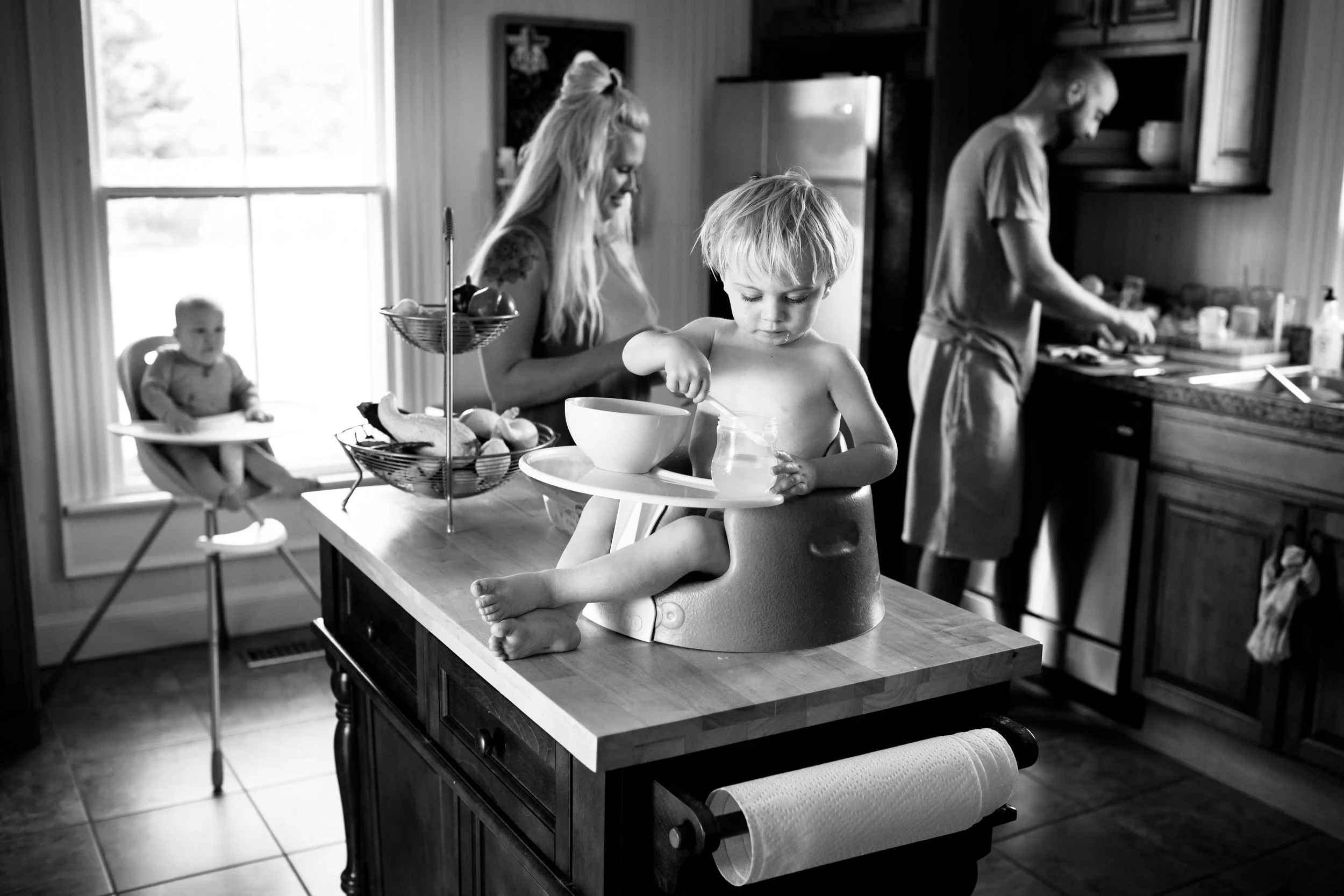 Black and white photo of a family in a kitchen. A woman is preparing food nearby a man who is at the stove. A young boy is sitting in a high chair at the table, and an older child is seated in a high chair, eating from a bowl.