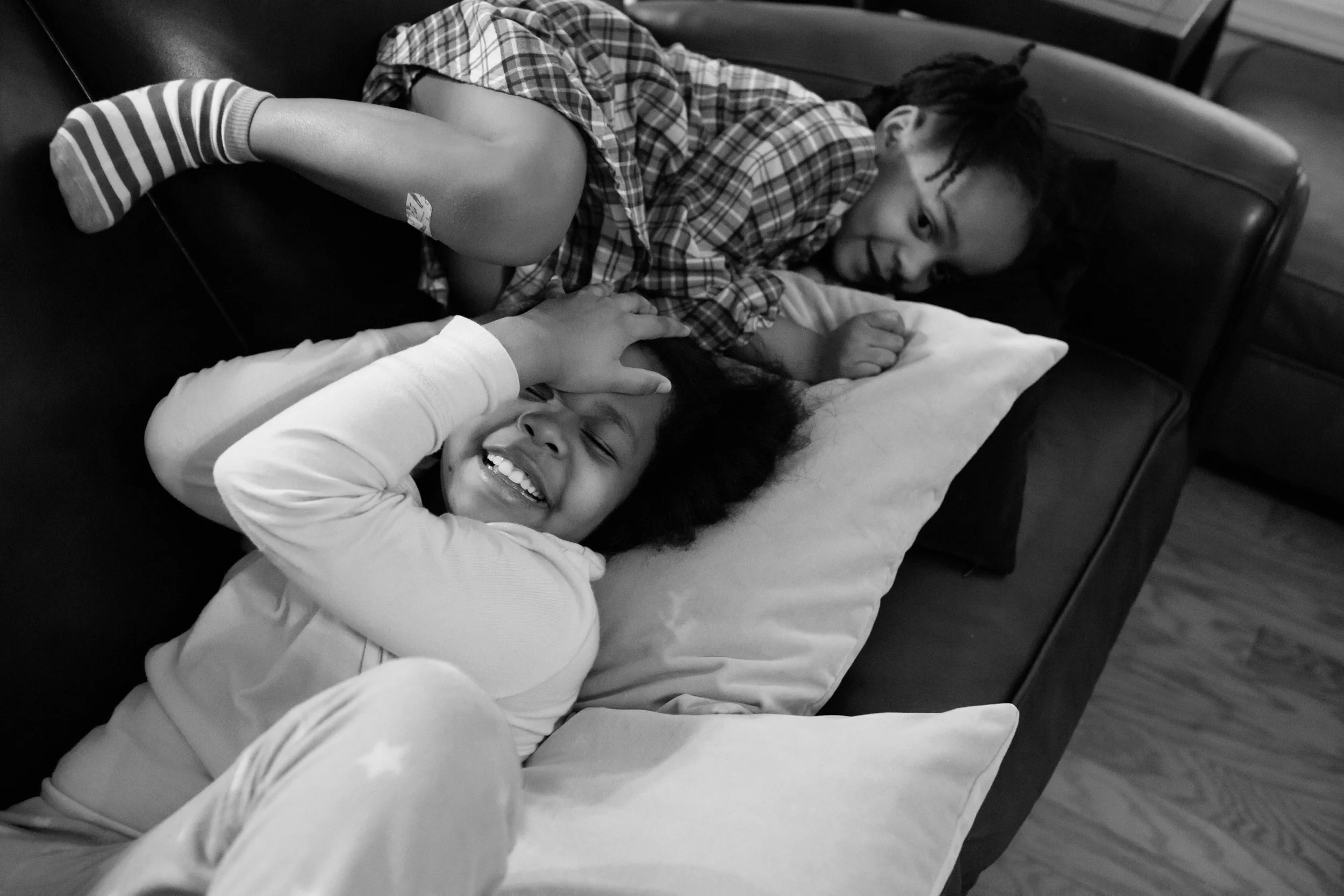 Two children, a girl with braids and a boy with an afro, lying on a hospital bed. The girl wears striped socks and pajamas, smiling and holding her head with one hand, while the boy is lying facing her with a big smile. An adult is gently holding the