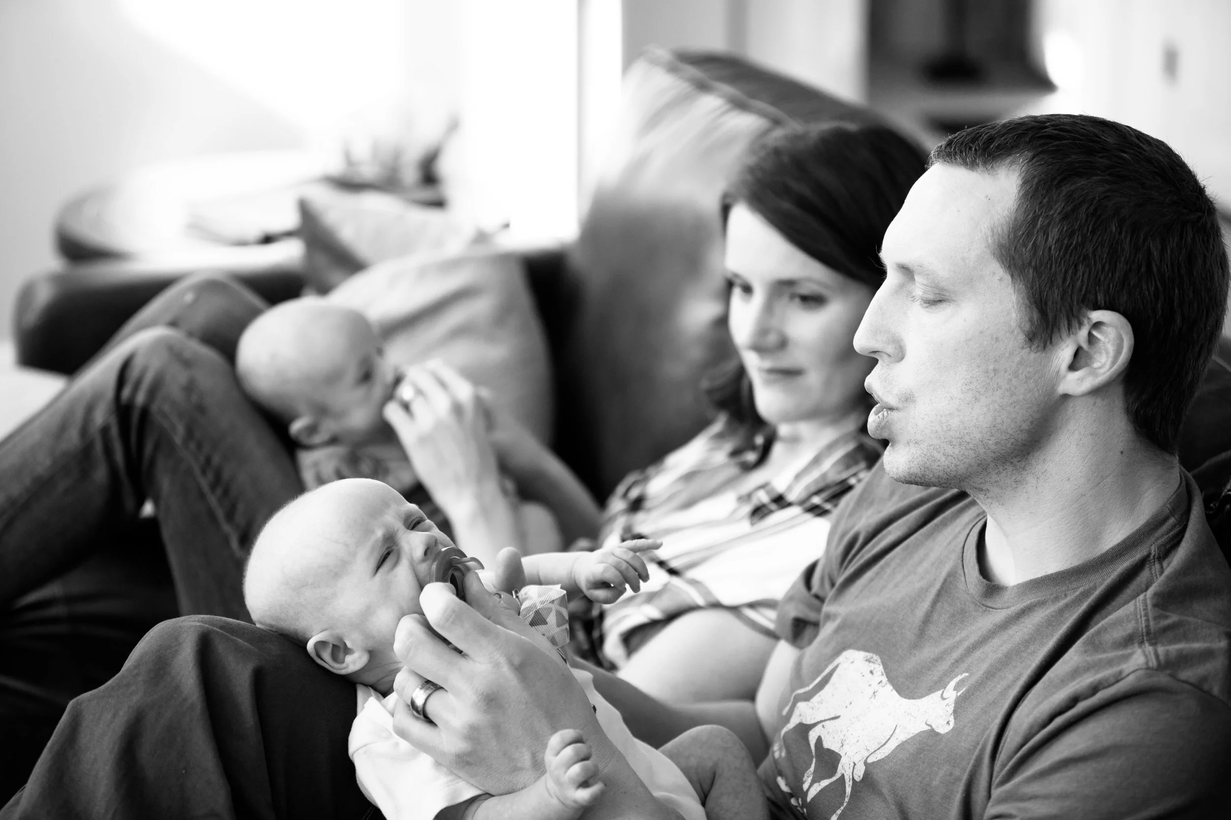 A black and white photo of a man and woman sitting on a couch, looking at a baby in the man's hands. The man is wearing a t-shirt and has short hair, while the woman has long hair and is looking at the baby. The baby is reaching out and holding a toy