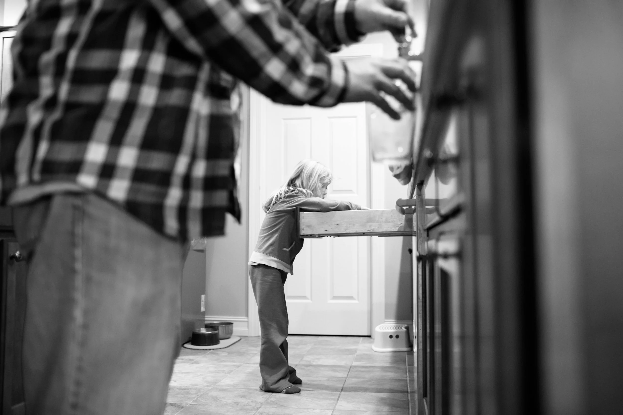 A young girl with long hair wearing a hoodie and sweatpants, standing in a kitchen, looking down at a wooden or plastic raised feeder or shelf. The photo is black and white and is taken from a low angle, with part of a countertop or furniture in the 