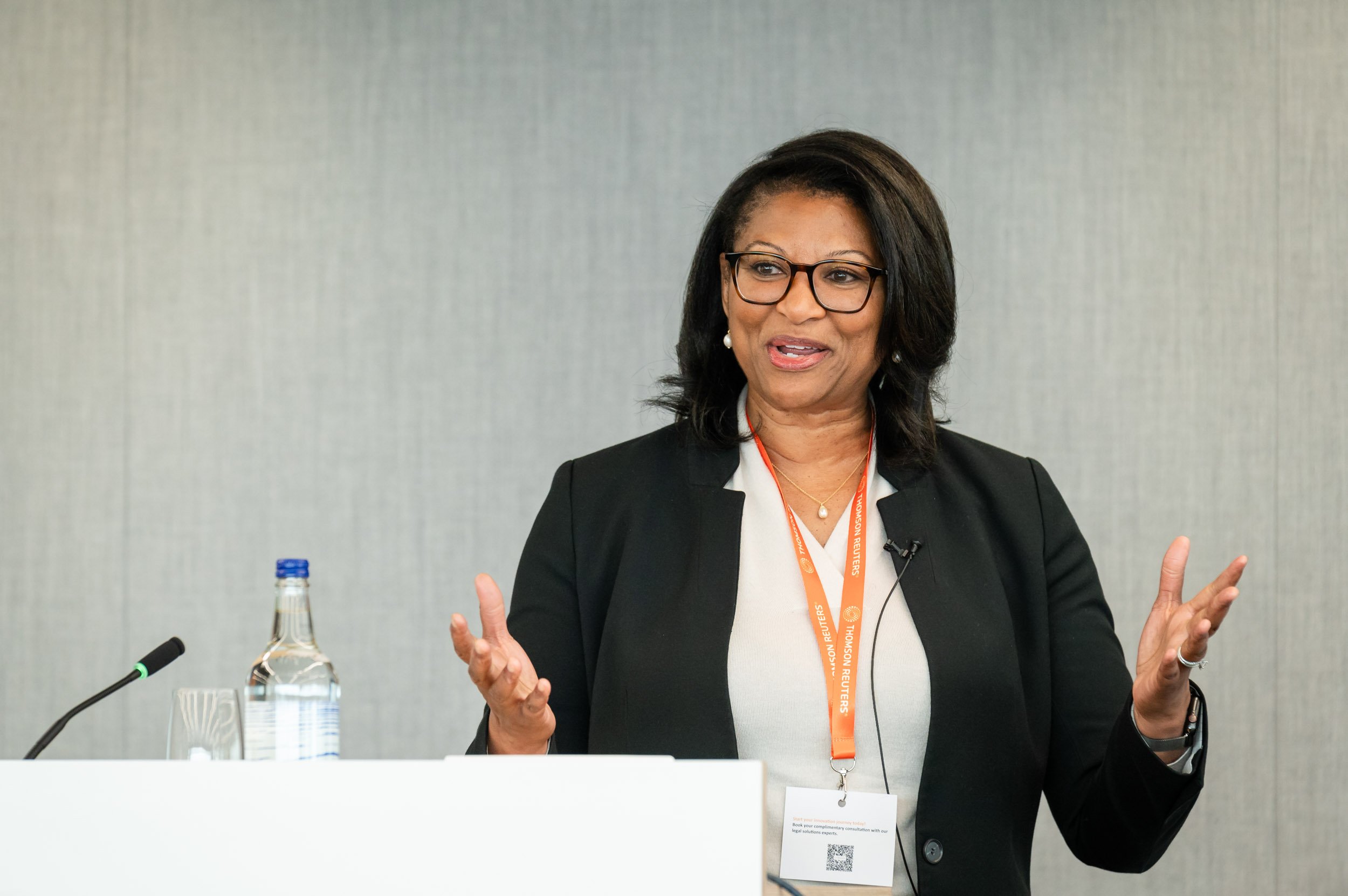 A professional event photograph of a keynote speaker at a podium in a prestigious London venue, capturing the professional scale and energy of a corporate summit.