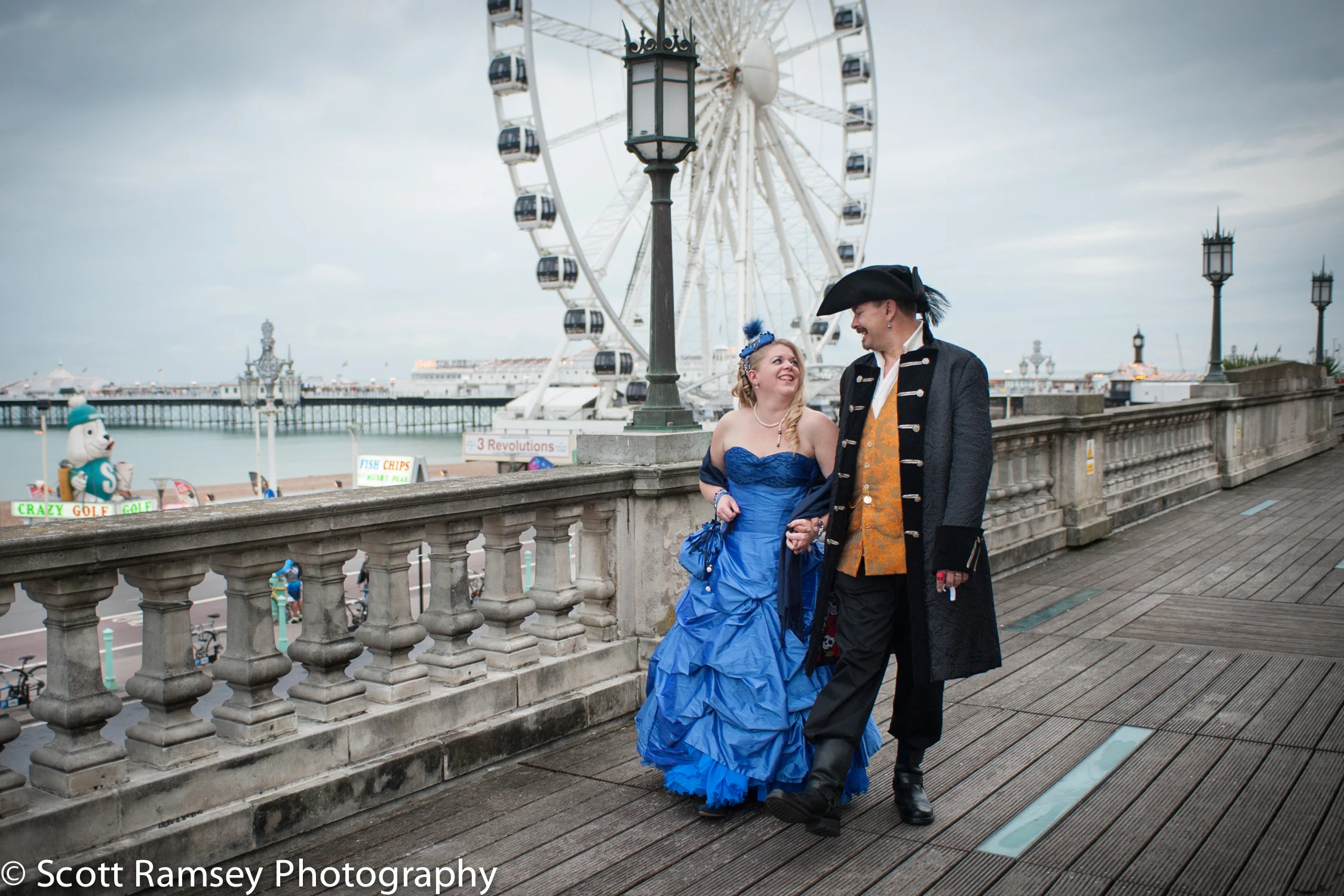Wedding Photography in Brighton. A very fun Pirate themed wedding in Brighton, East Sussex. In Pic; The Bride and Groom (Dressed as a Pirate) walk hand in hand past the wheel on the seafront in Brighton.