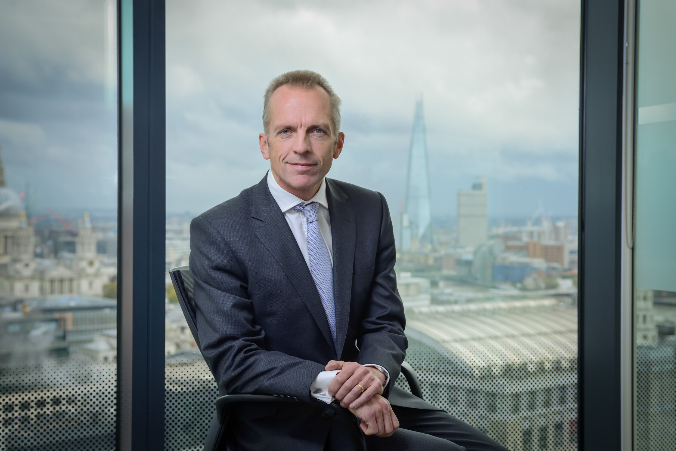 A confident and professional corporate headshot of a male CEO seated in a London office, featuring a cinematic view of the city skyline through the window as a natural office environment.
