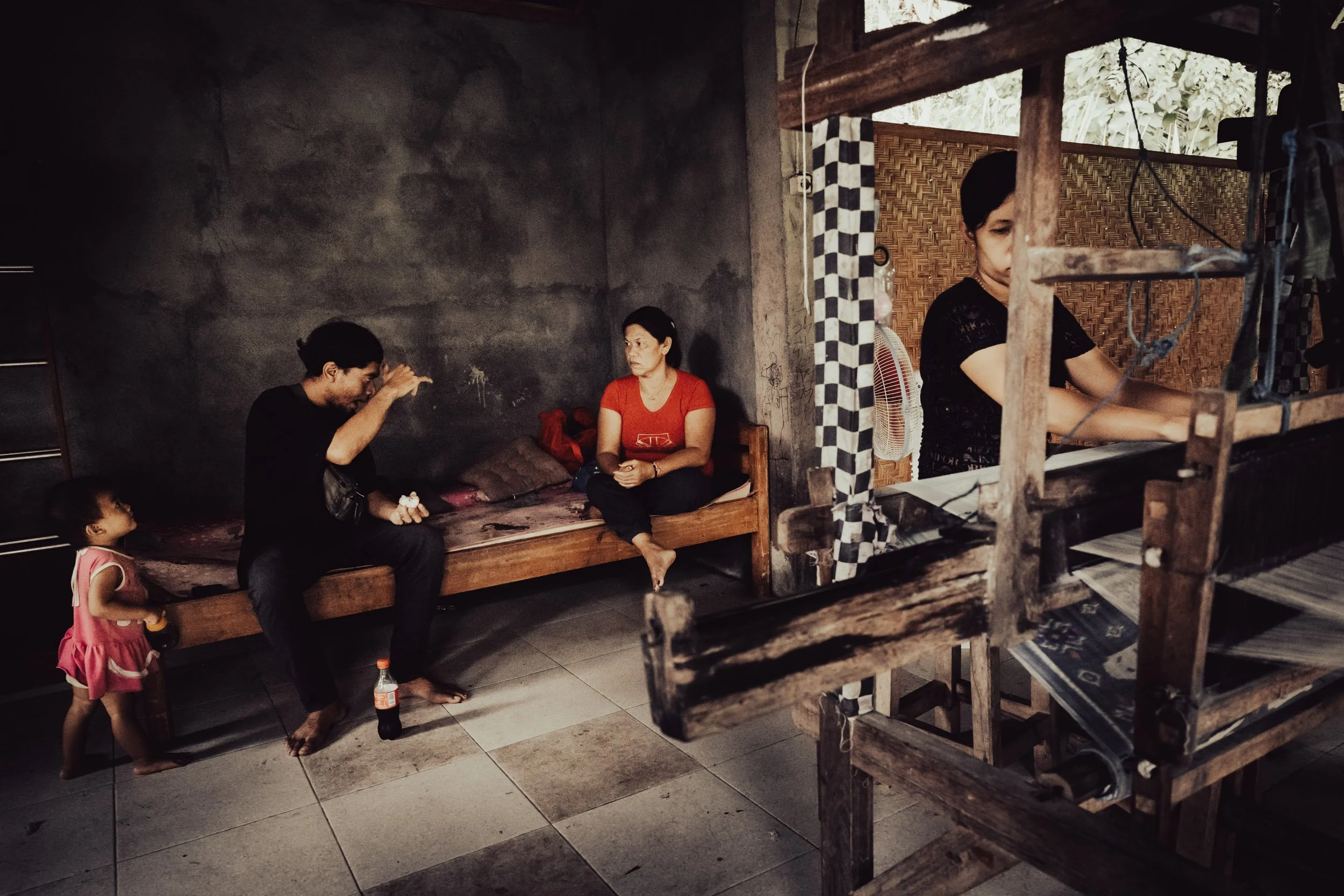 Documentary photograph in Bengkala village, Bali, featuring two individuals communicating via Kata Kolok sign language beside a woman operating a traditional weaving loom, with a young child observing the scene.