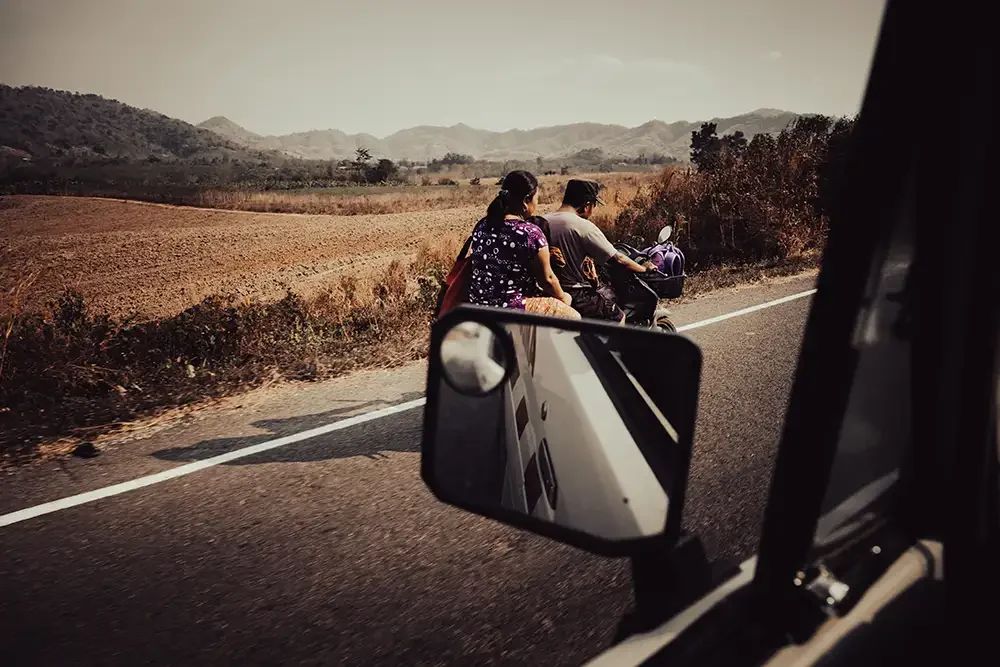 A documentary photograph taken through an ambulance window showing a family of three on a motorcycle traveling near the Thai-Myanmar border.