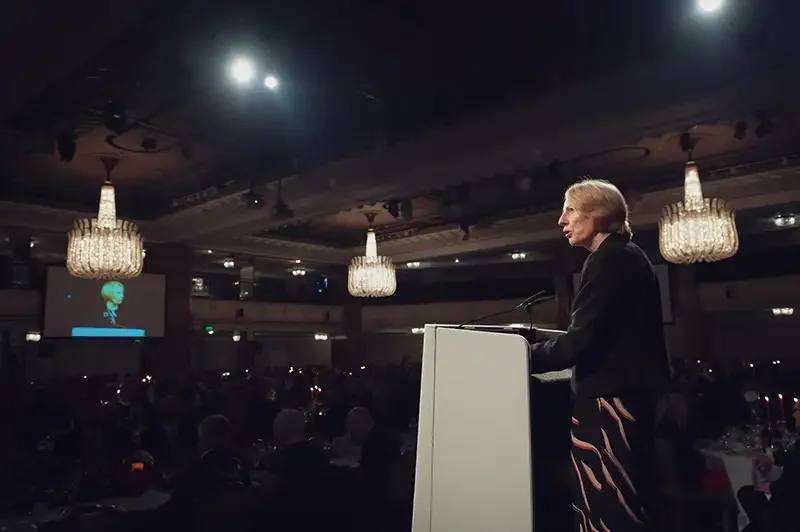 Atmospheric wide shot of a female keynote speaker addressing a conference at a luxury London hotel. Editorial corporate event photography capturing the scale and lighting of the ballroom.