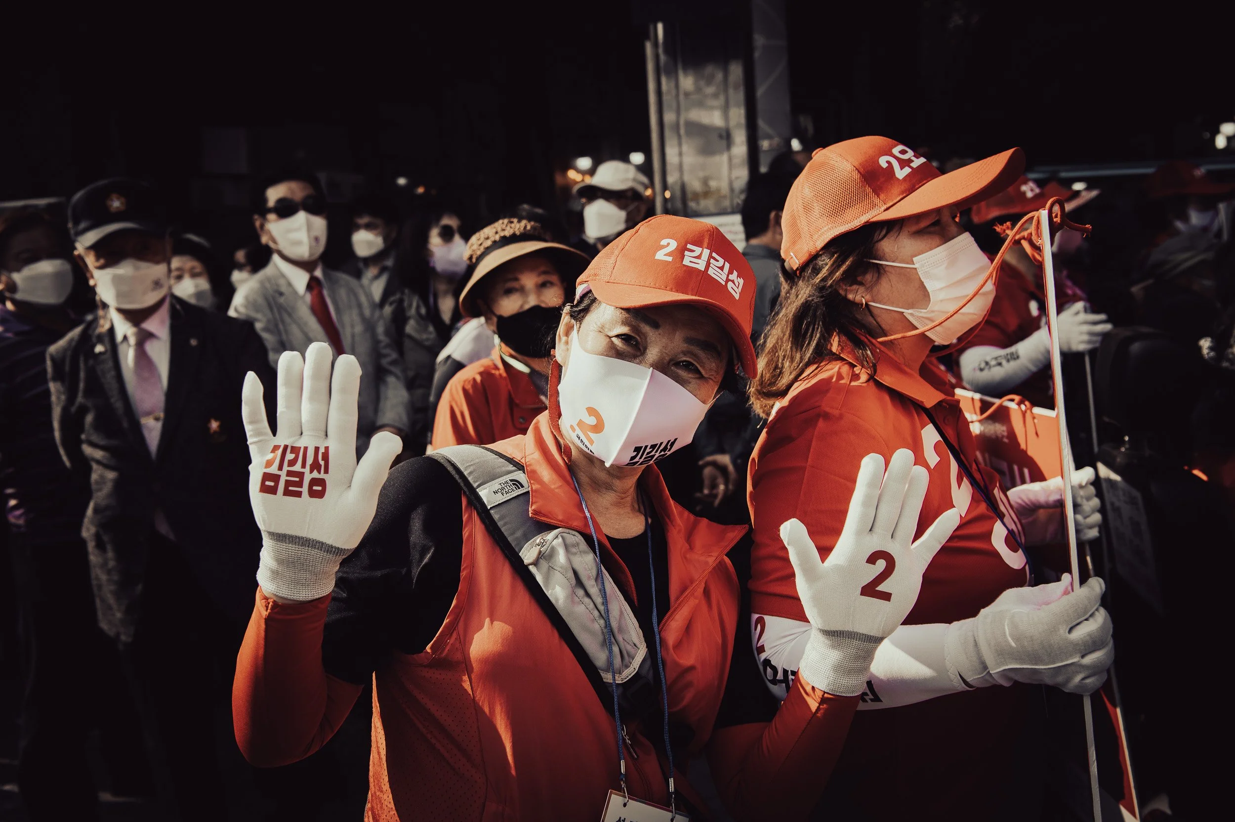 Senior woman at a Seoul Central Market political rally wearing red with campaign badges and raising white-gloved hands. South Korea street photography by Scott Ramsey.