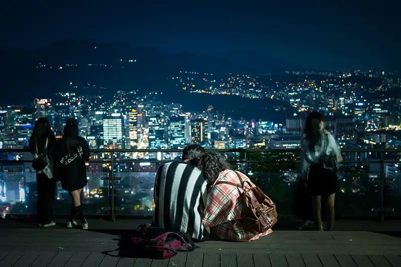 A young couple sits together at the Namsan Tower (N Seoul Tower), overlooking the vibrant night skyline of Seoul. Captured by Scott Ramsey, this image illustrates the intersection of ancient spirit and modern resilience in South Korea.