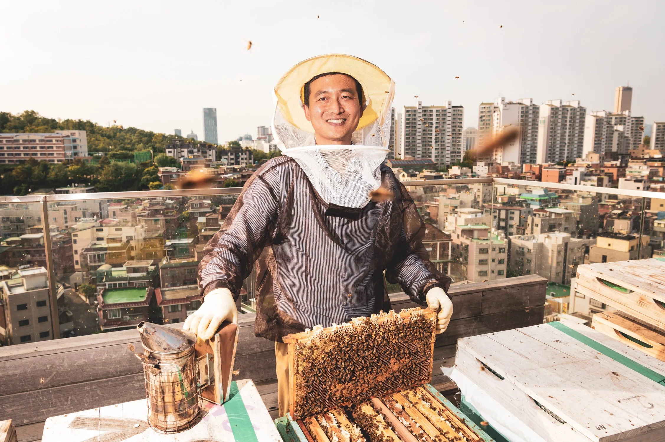 An urban beekeeper tending to hives on a hotel rooftop overlooking the city skyline of Seoul, South Korea, captured by documentary photographer Scott Ramsey.