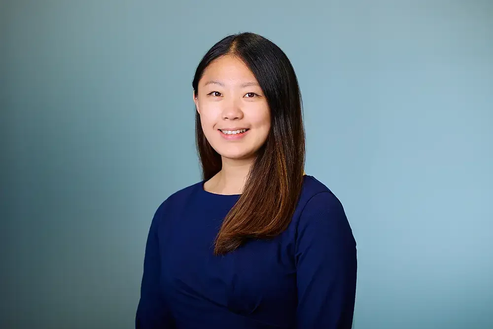 A professional corporate headshot of an approachable female employee, photographed against a mobile backdrop inside a London office.