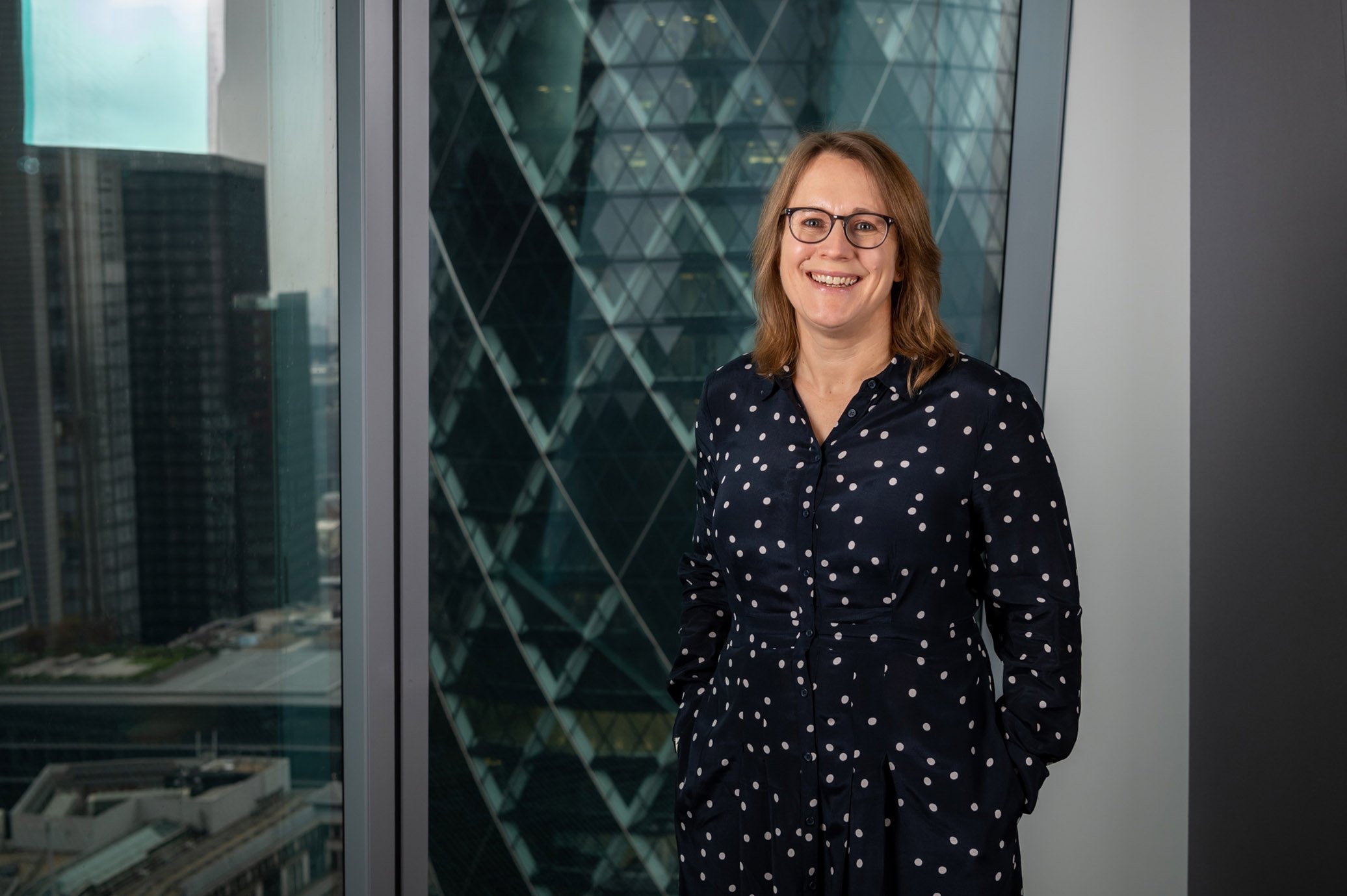 Wide-angle editorial corporate portrait of a woman in a blue dress standing in front of a window on the 20th floor, with an overcast, cinematic November view of The Gherkin and the London city skyline.