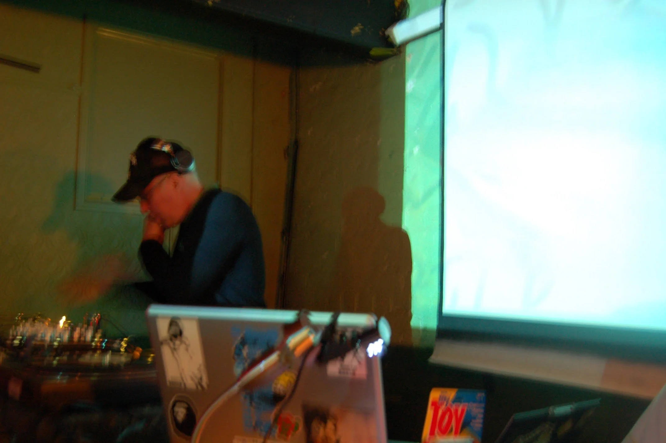 A person with a black cap and navy blue shirt DJing with turntables in a dimly lit room, with a neon green screen or projection on the wall.