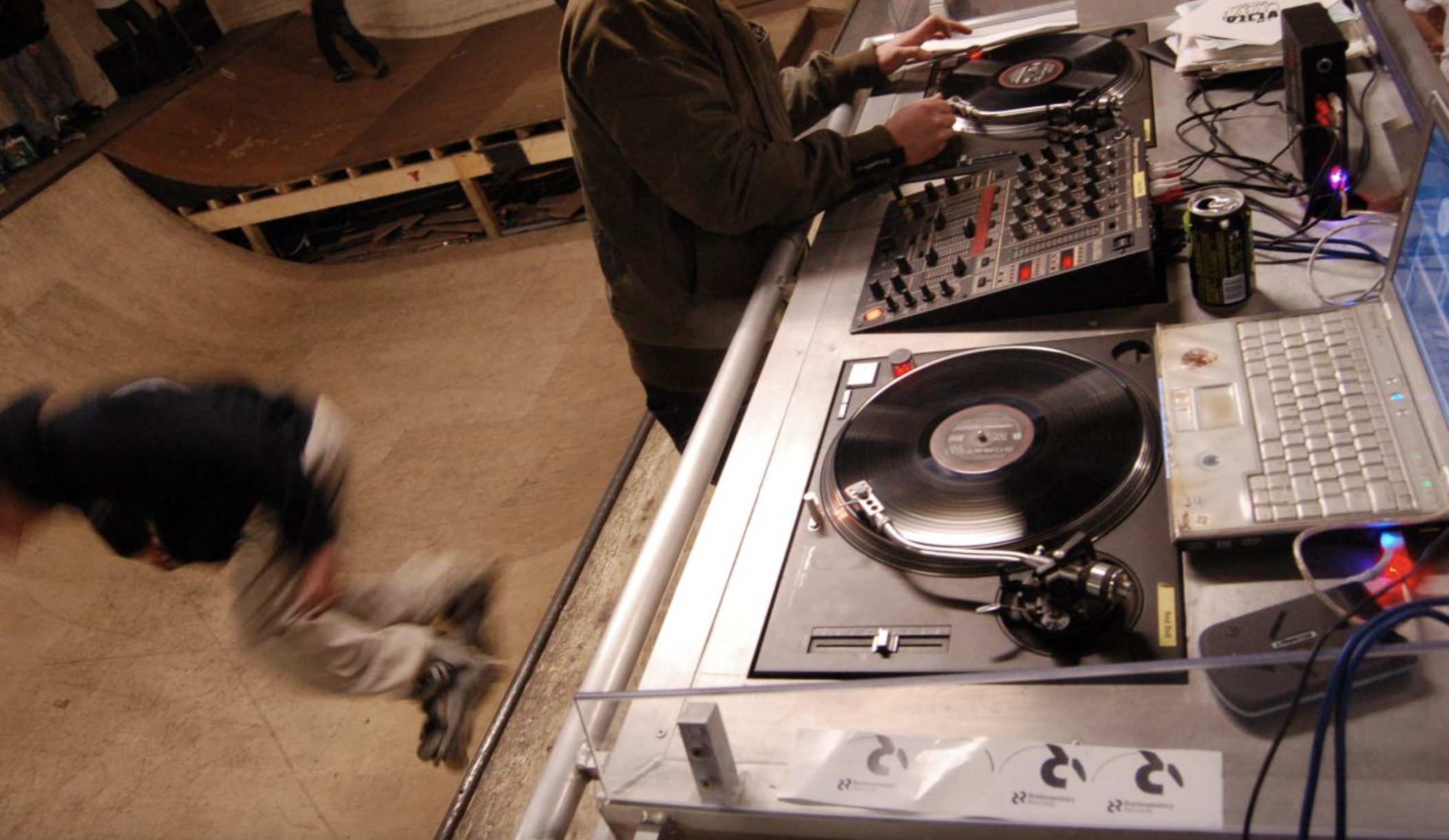 A DJ mixing music with turntables, a mixer, and a laptop on a table in a room with a curved ramp and a person standing on the ramp.