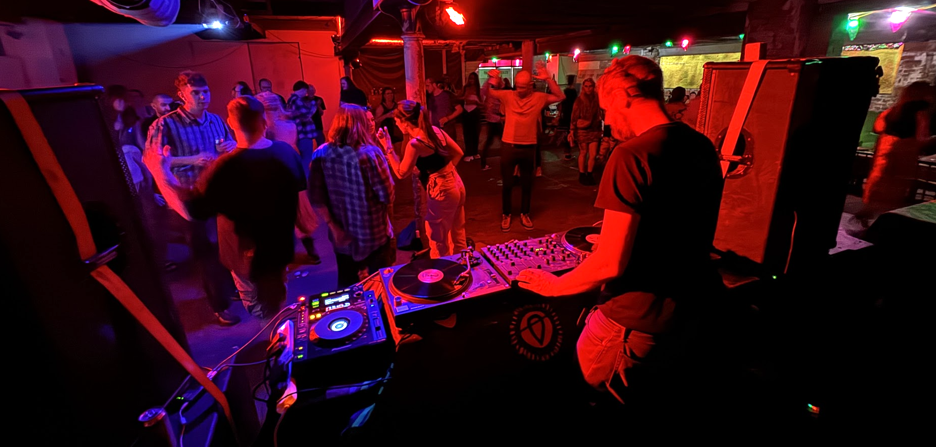 DJ performing at a nightclub with people dancing and socializing in a dimly lit room with colorful lights.