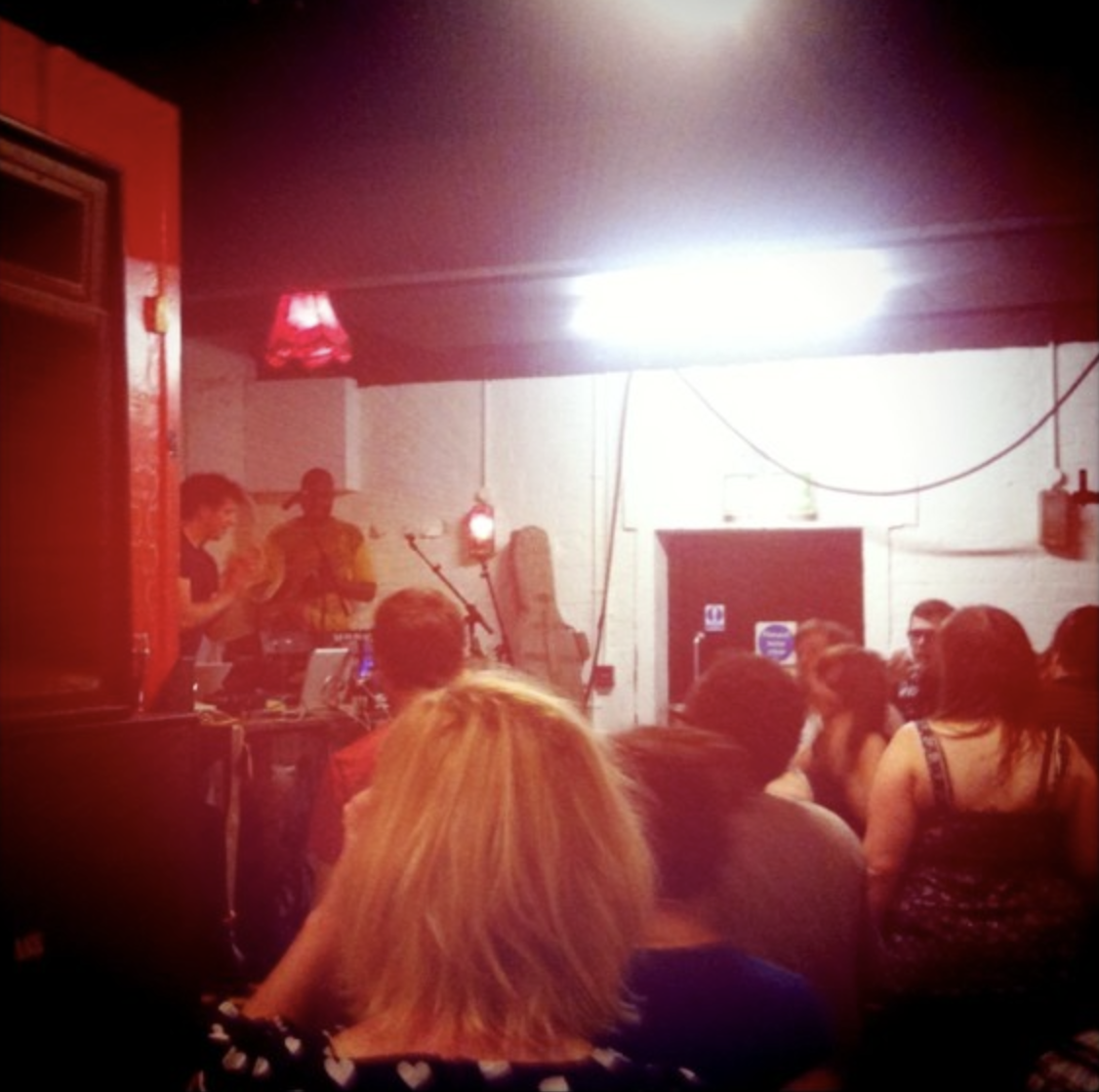 A live music performance in a small indoor venue with a performer on stage, surrounded by an audience seated and standing, illuminated by low lighting and red ambiance.