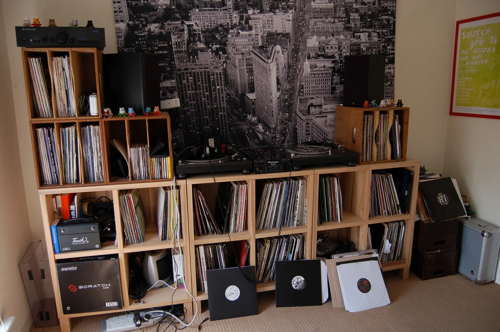 A room corner with wooden shelving holding vinyl records, a turntable, and gaming equipment. A black and white cityscape wall mural is in the background, and a framed poster is on the right wall.