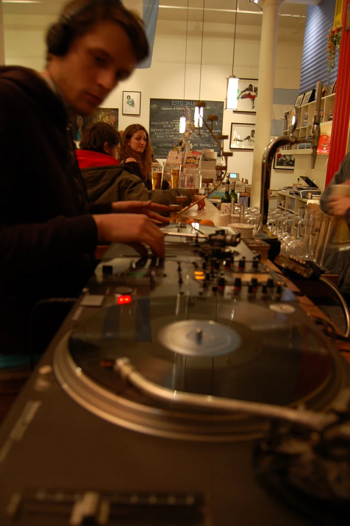 A person DJing with a turntable in a lively cafe or bar with other patrons, artwork on the walls, and a chalkboard menu in the background.
