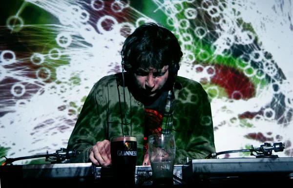 A DJ with headphones standing behind a turntable, with a Guinness pint glass and a glass of water in front, in front of a colorful abstract background.