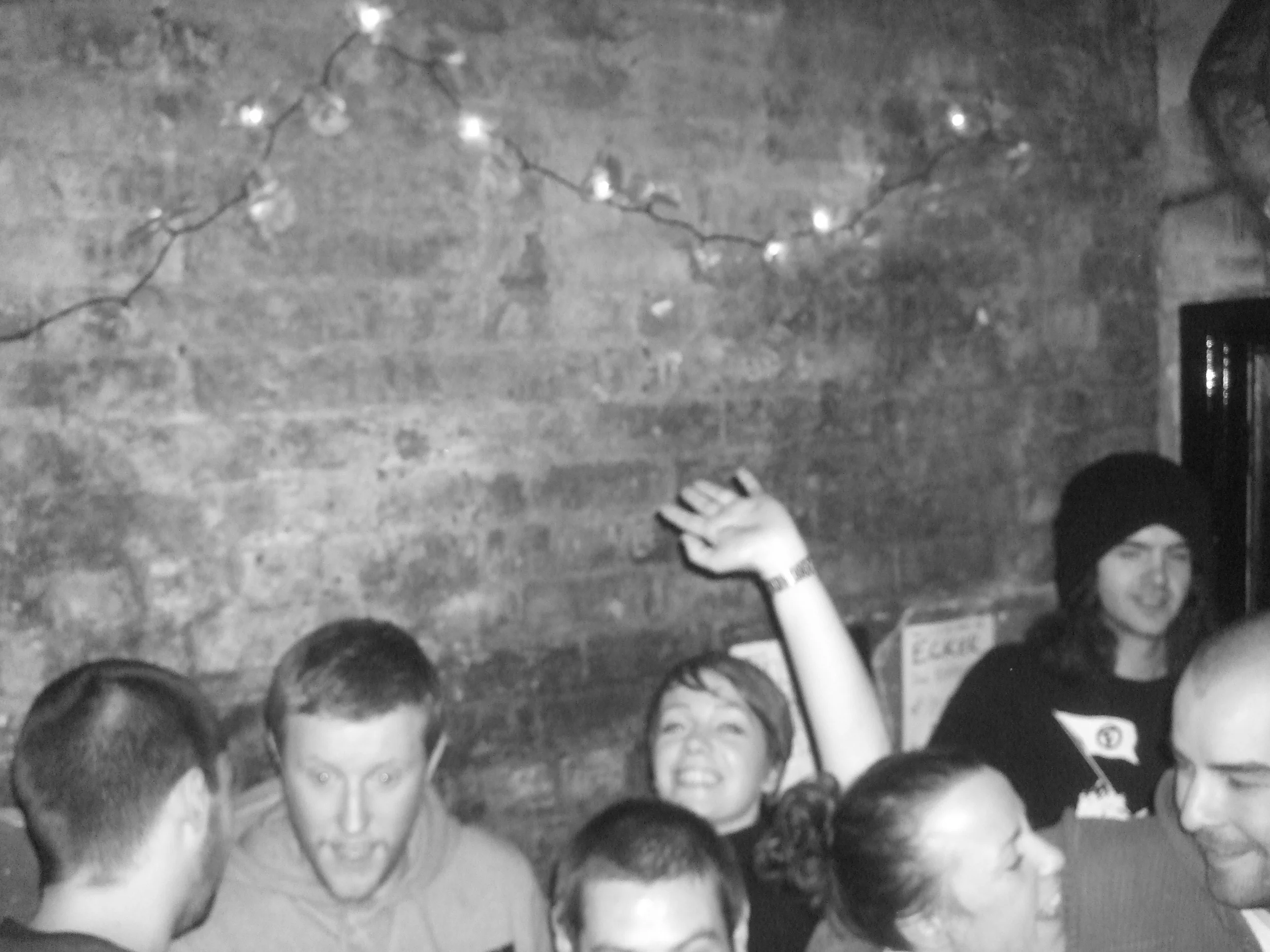 Group of people socializing with a brick wall background and string lights overhead, one person raising their arm and smiling.