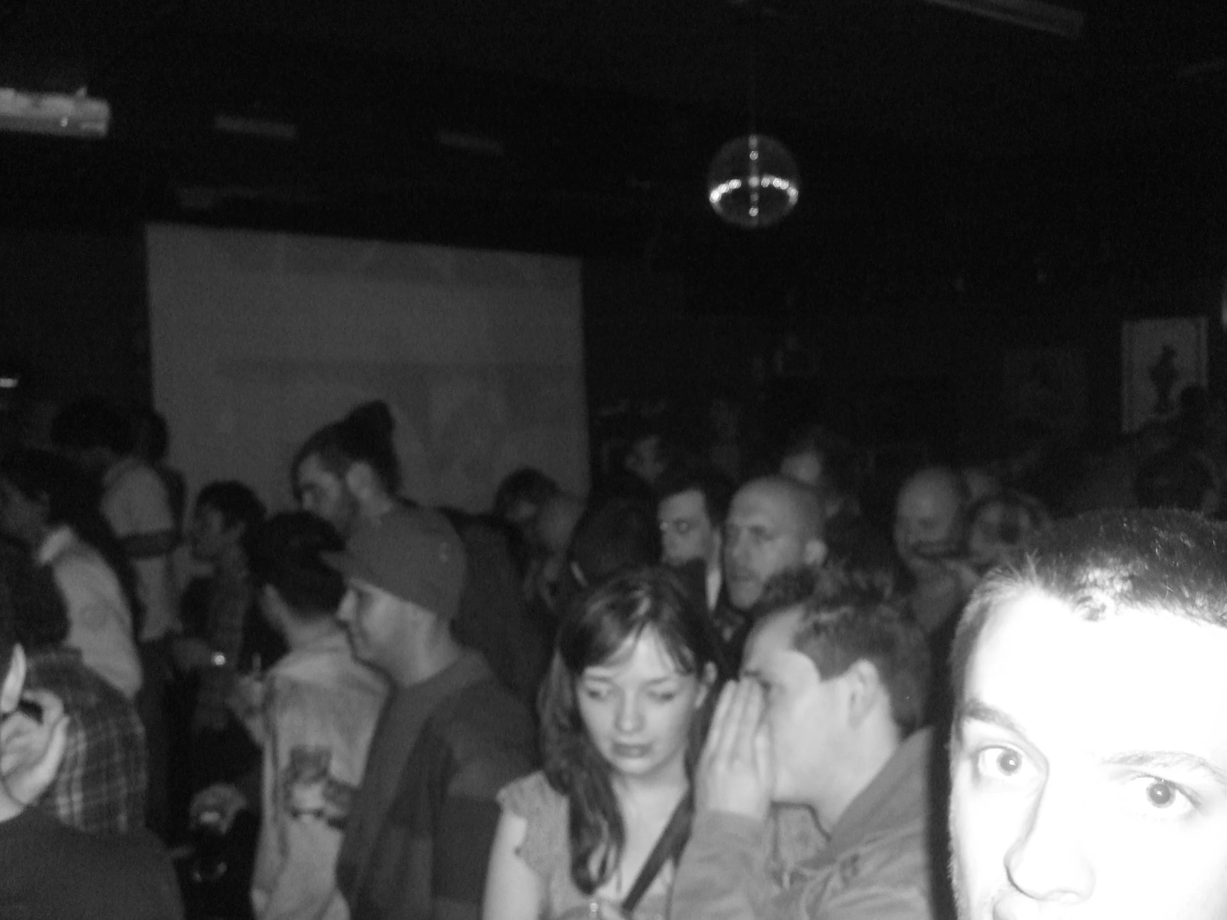 A crowded indoor party or concert with people standing close together, some talking or looking around, and a disco ball hanging from the ceiling.