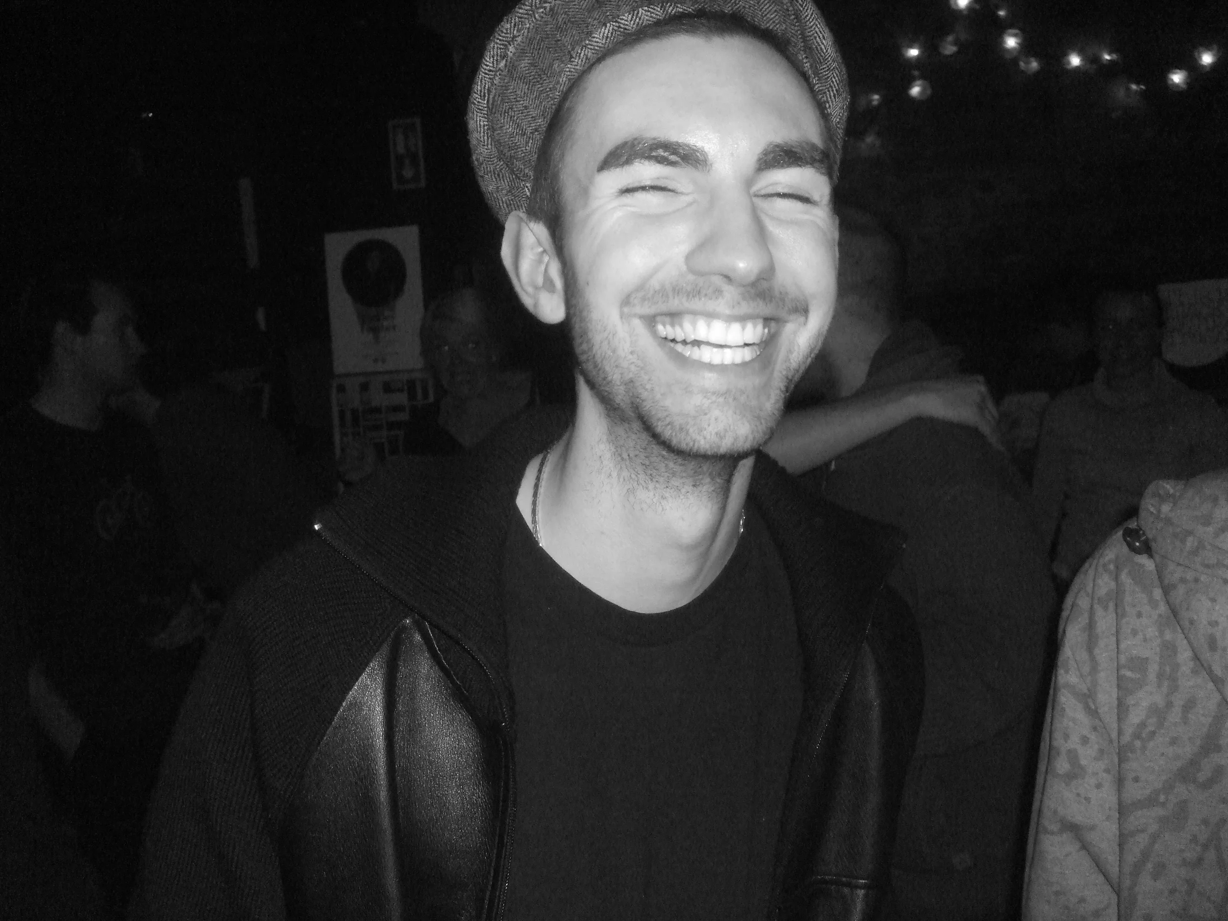 A smiling young man wearing a beanie and black leather jacket in a dimly lit social gathering, with other people in the background.