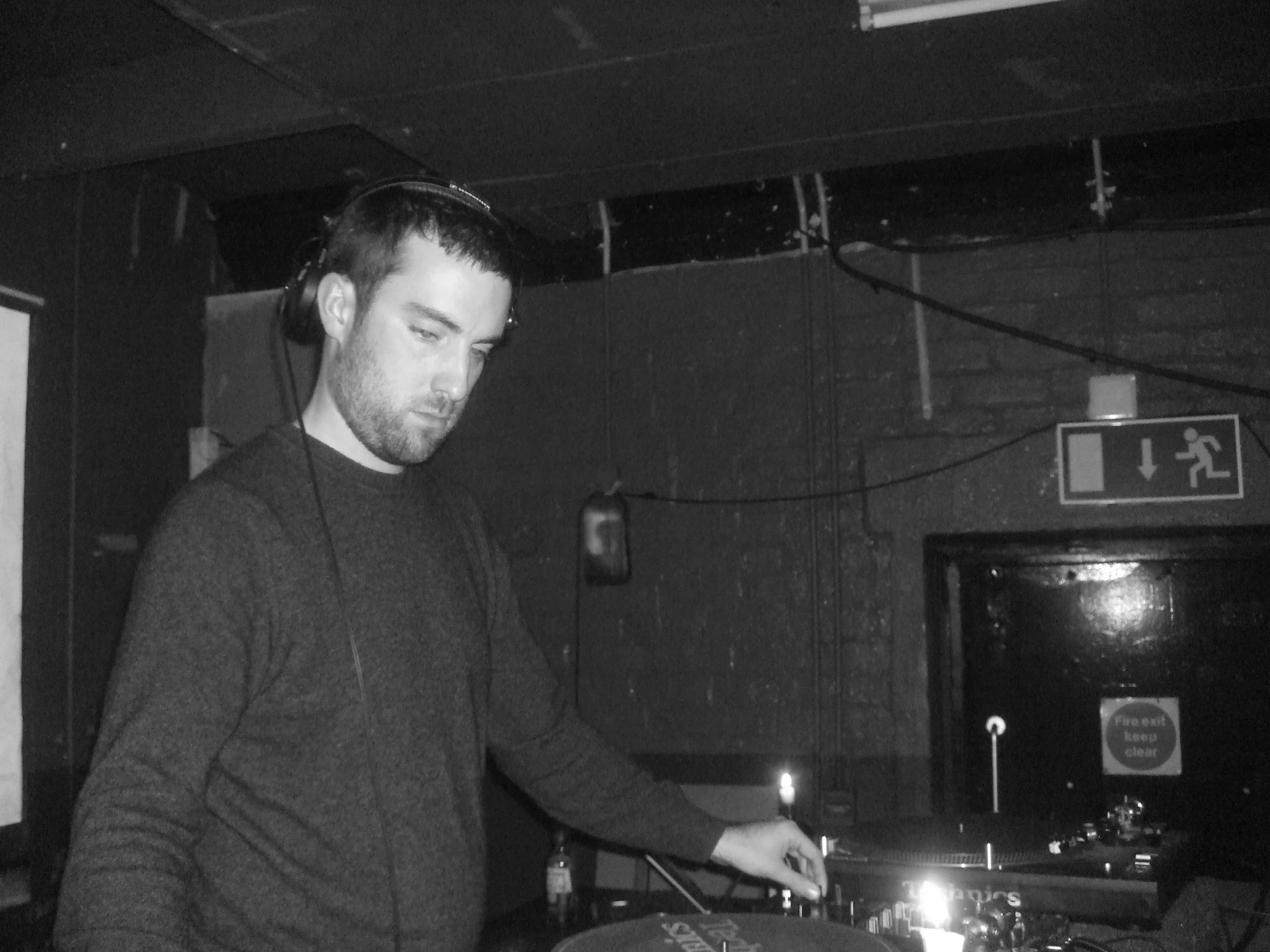 A man with headphones DJing at a club or event, with a dark background and emergency exit sign overhead.