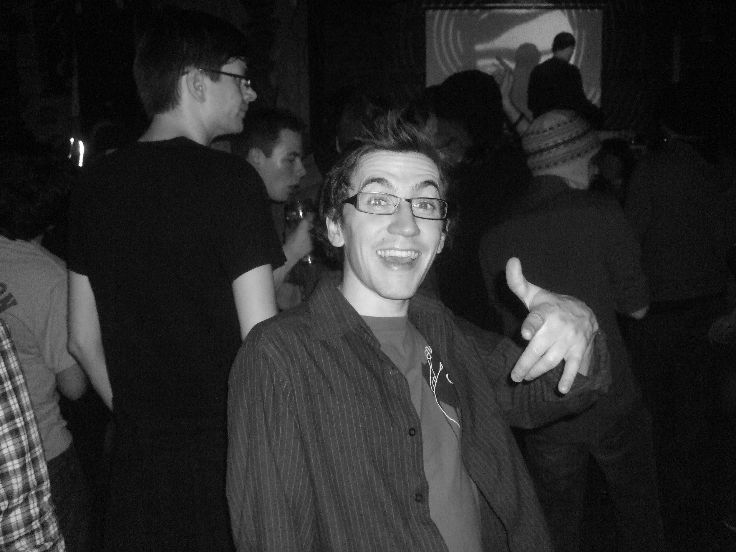 A man smiling and showing a peace sign at a crowded event or party, with other people in the background.