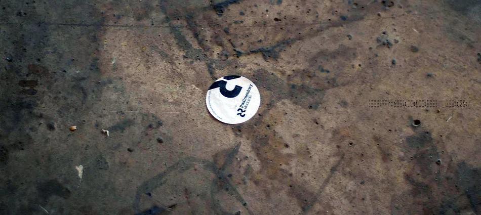 Close-up of a dirty, dusty surface with a small round sticker or token featuring a black number '2' and text 'Platinum Currency'.