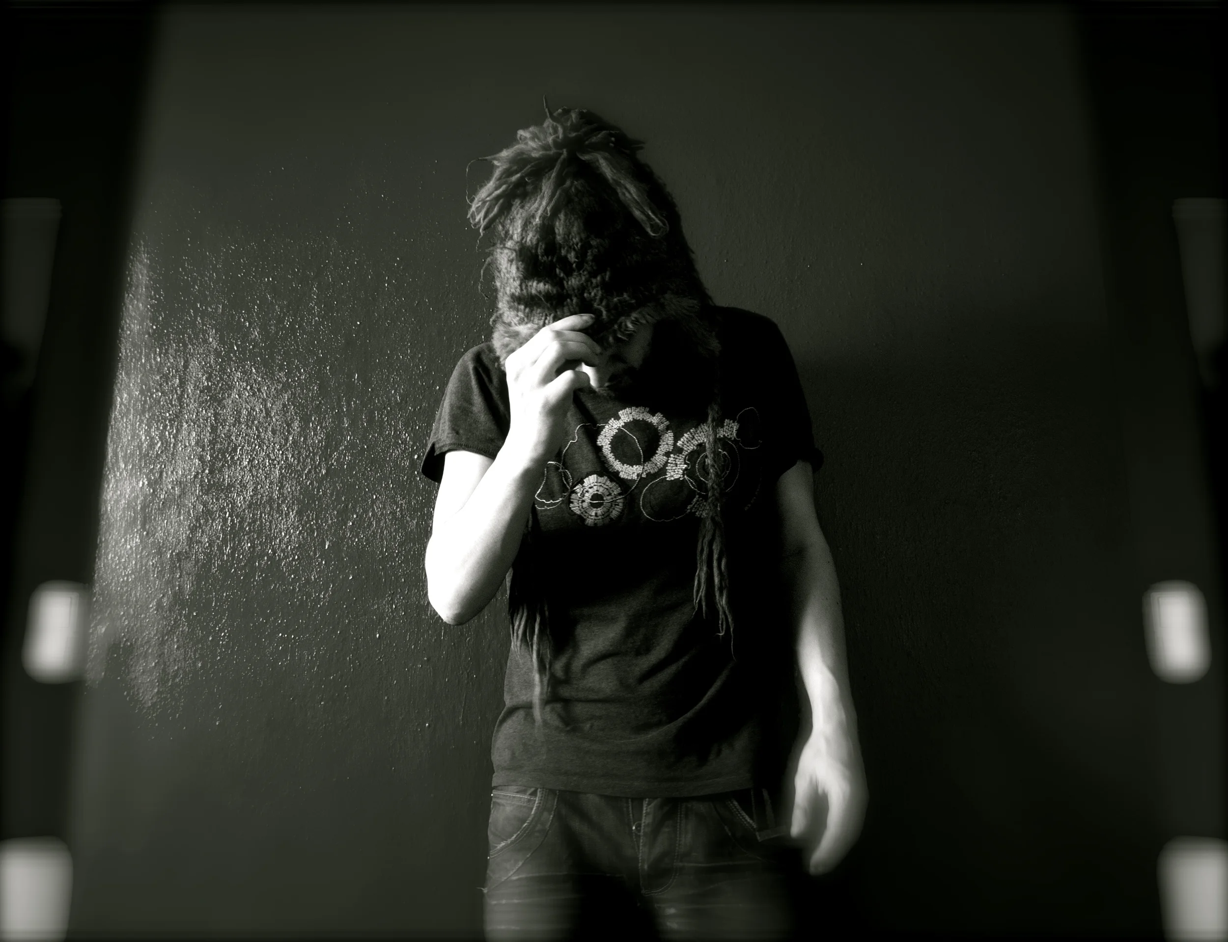 Young person with dreadlocks standing against a dark wall, head bowed, hand on forehead, wearing a black t-shirt with abstract design.
