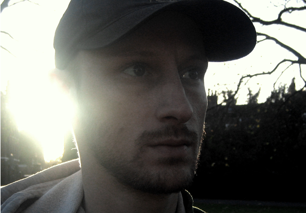 A man in a baseball cap standing outdoors with the sun setting behind him, creating a backlit silhouette.